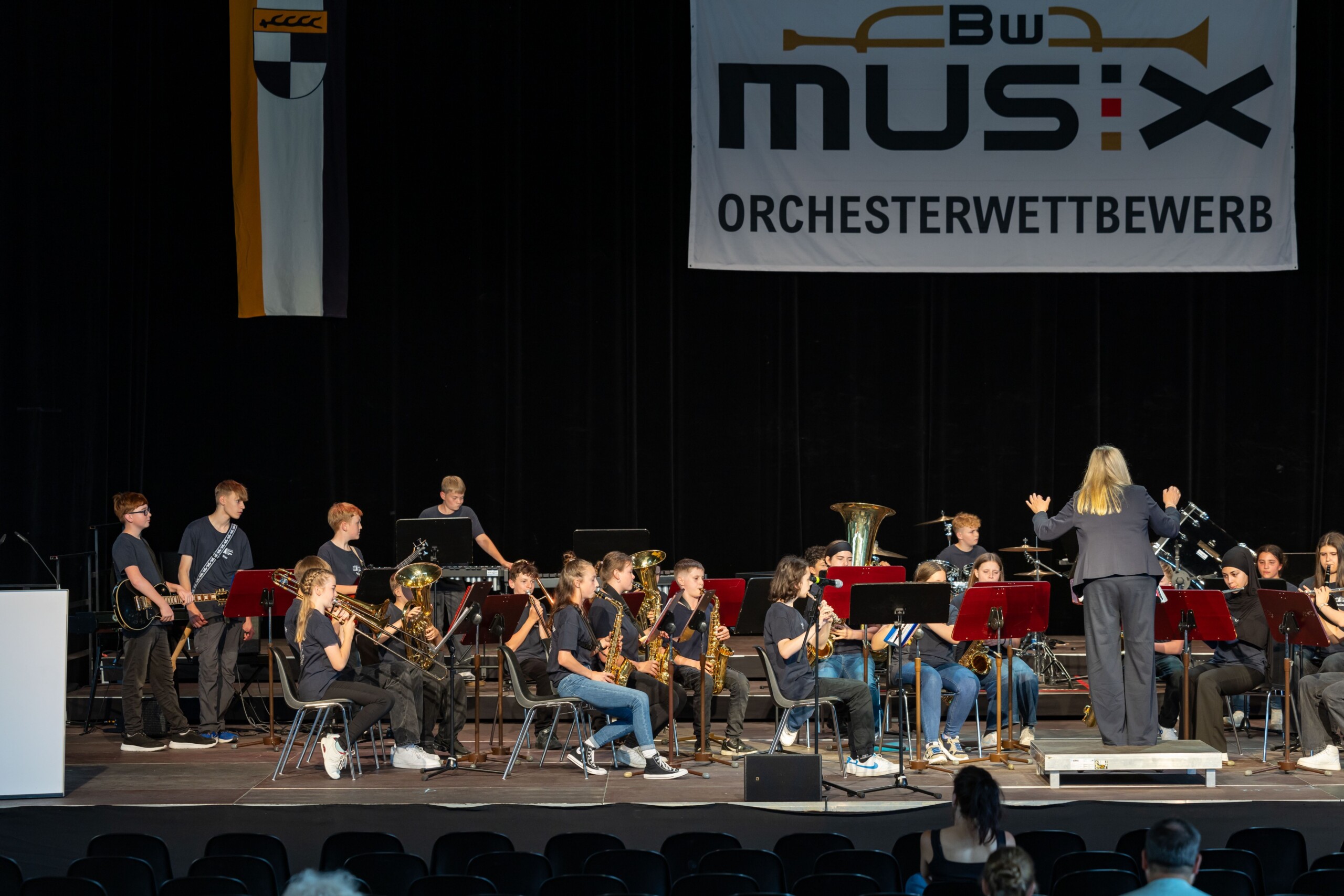 Gesamtaufnahme der Bühne mit der Bläserklasse 7d beim BW Musix-Wettbewerb – das gesamte Orchester ist in Aktion, begleitet von intensiver Dirigierarbeit durch Christine Jahn.