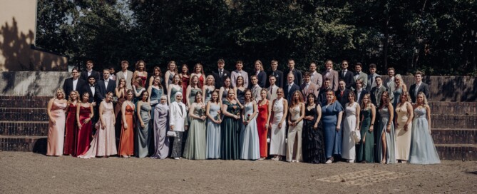 Gruppenfoto des Abiturjahrgangs 2025 der Gesamtschule Waldbröl auf dem Schulhof – die Abiturient:innen stehen festlich gekleidet in mehreren Reihen vor einer grünen Baumkulisse und lächeln in die Kamera.