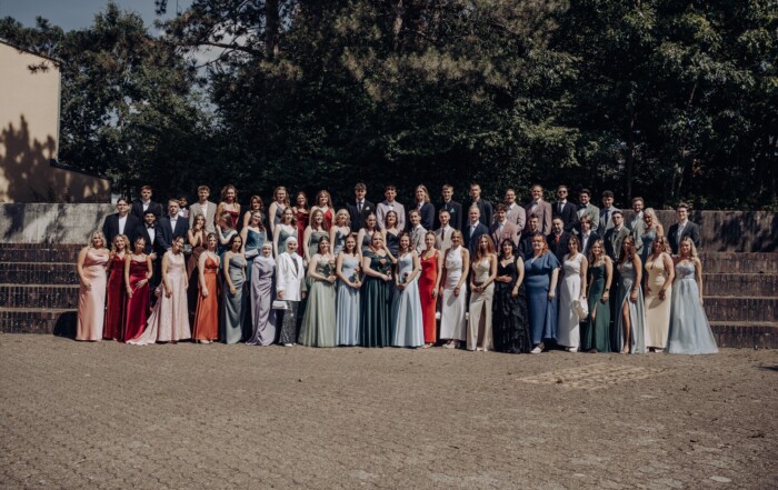Gruppenfoto des Abiturjahrgangs 2025 der Gesamtschule Waldbröl auf dem Schulhof – die Abiturient:innen stehen festlich gekleidet in mehreren Reihen vor einer grünen Baumkulisse und lächeln in die Kamera.
