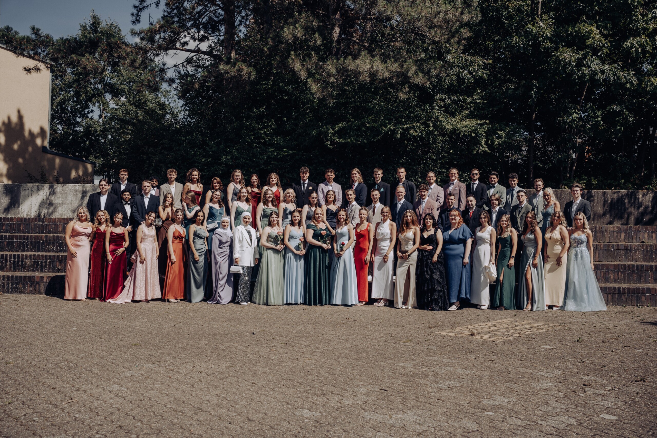 Gruppenfoto des Abiturjahrgangs 2025 der Gesamtschule Waldbröl auf dem Schulhof – die Abiturient:innen stehen festlich gekleidet in mehreren Reihen vor einer grünen Baumkulisse und lächeln in die Kamera.