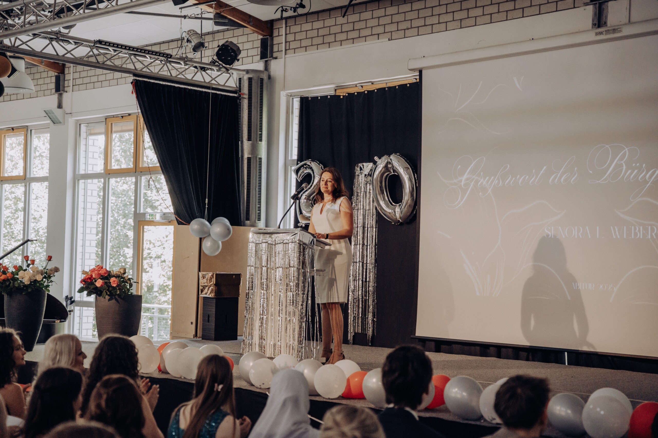 Bürgermeisterin Larissa Weber steht am Rednerpult der festlich geschmückten Mensa und hält ihr Grußwort – im Hintergrund große silberne Zahlenballons „20“ und „25“.