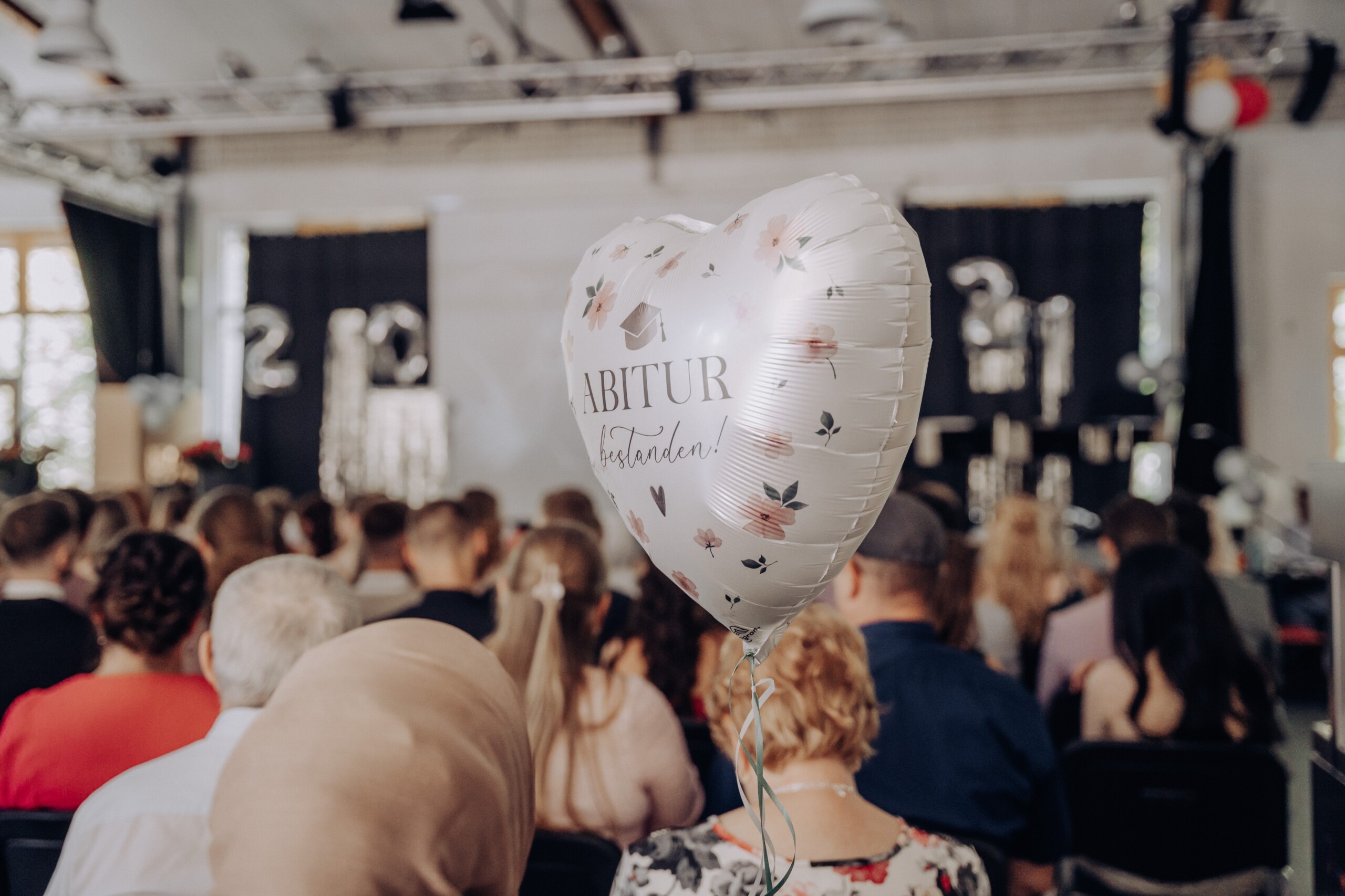 Ein herzförmiger Luftballon mit der Aufschrift „Abitur bestanden!“ schwebt zwischen den Zuschauer:innen und verleiht der Feier eine festliche, emotionale Atmosphäre.