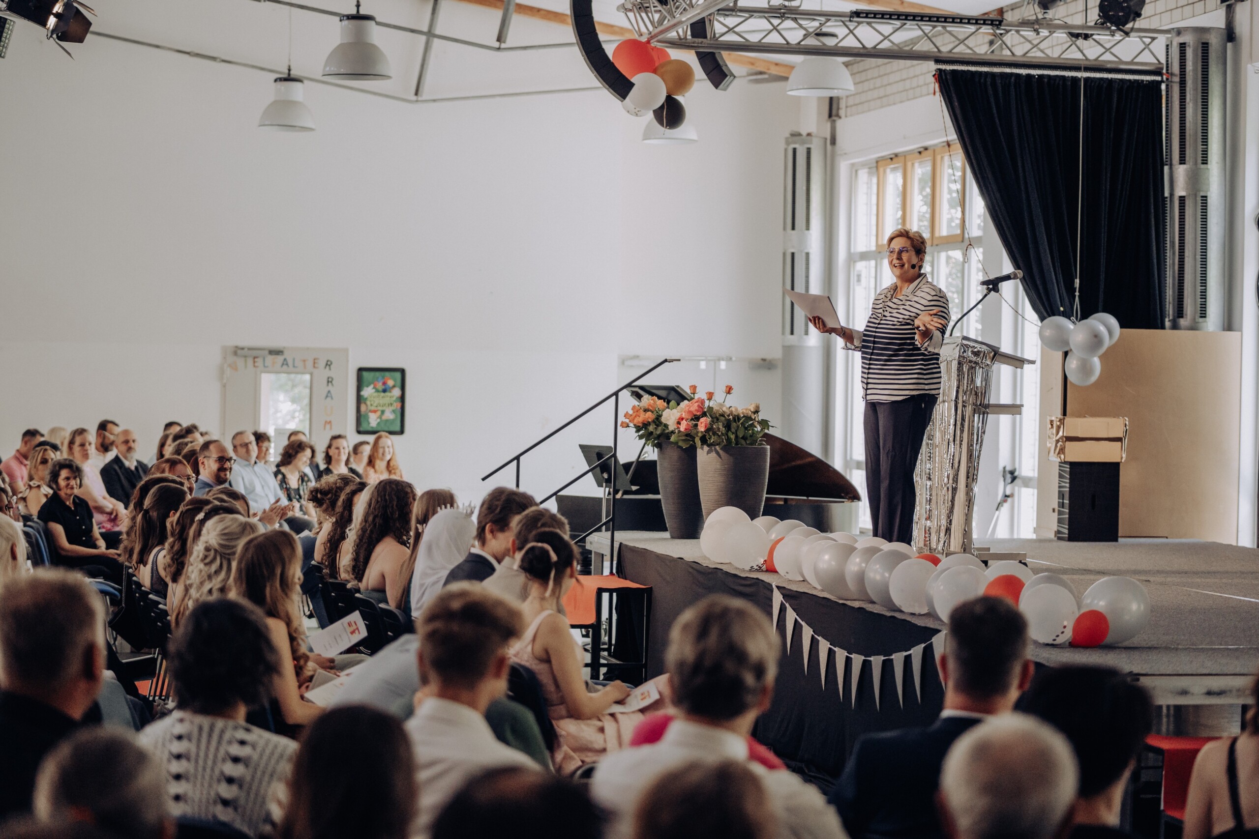 Oberstufenleiterin Ute Gick spricht mit einem Lächeln zu den versammelten Gästen – im Vordergrund sind Schüler:innen und Eltern zu sehen, die aufmerksam zuhören.