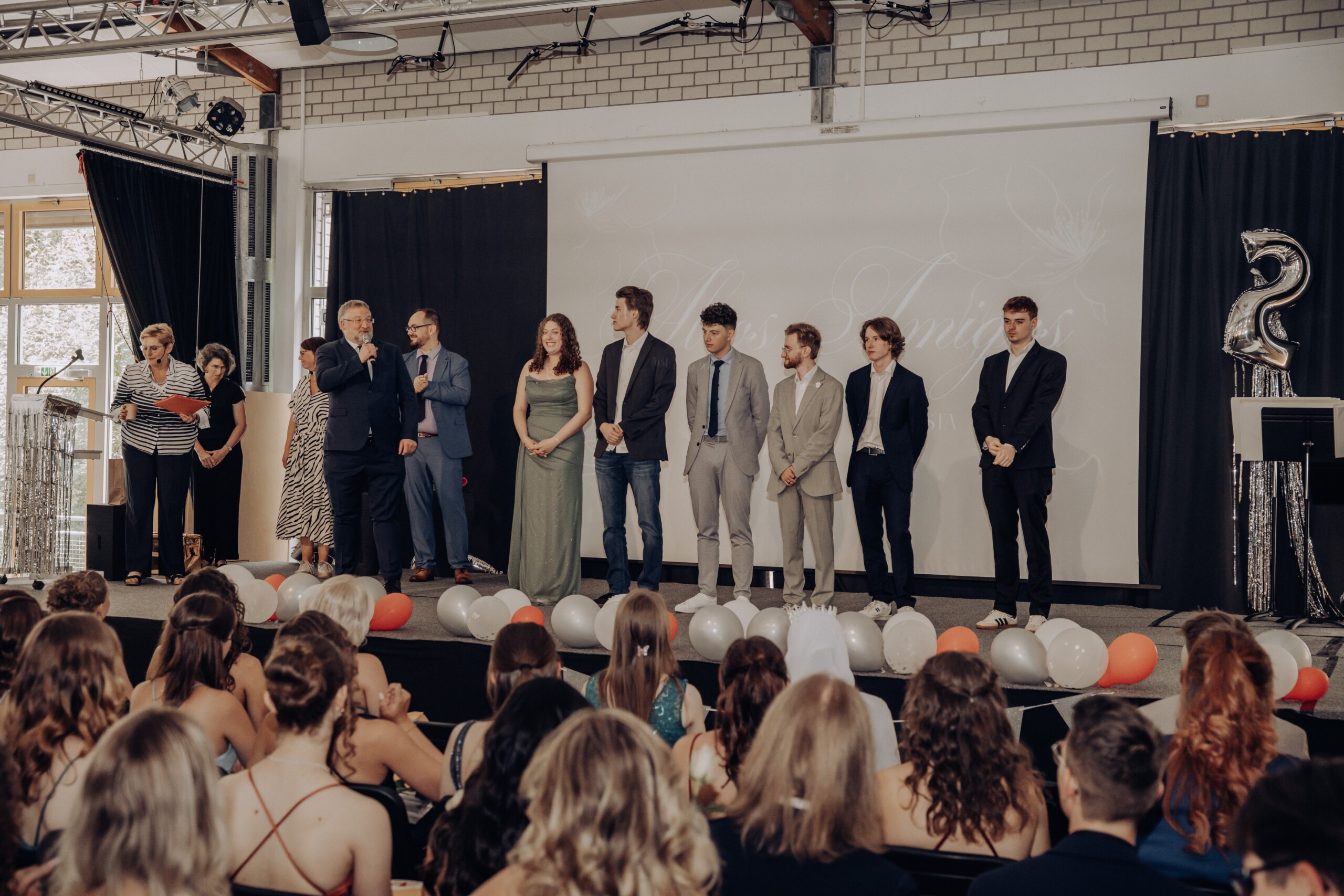 Die sieben Latinum-Absolvent:innen stehen auf der Bühne, begleitet von Lehrkräften – im Vordergrund verfolgen Mitschüler:innen und Gäste das Geschehen.