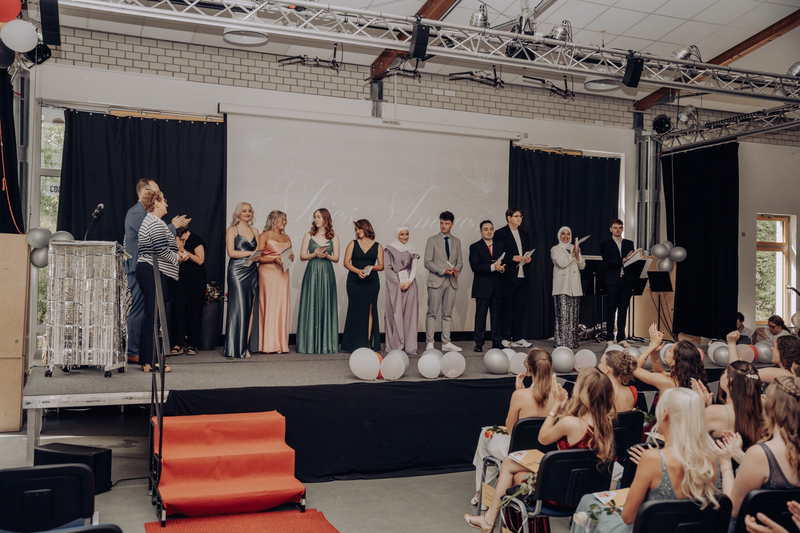 Zehn Abiturient:innen werden auf der Bühne für ihre hervorragenden Leistungen mit einer Eins vor dem Komma geehrt – Applaus aus dem Publikum begleitet die Ehrung.