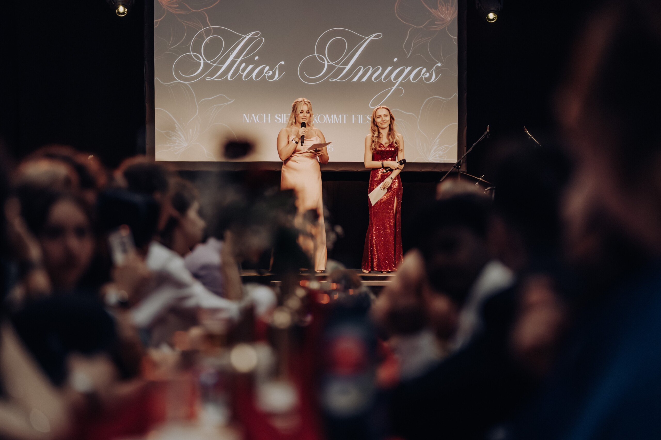 Walentina Krischke und Sarah Bauermann führen durch das Bühnenprogramm des Abiballs – im Hintergrund das Motto „Abios Amigos – Nach Siesta kommt Fiesta“ auf einer großen Leinwand.