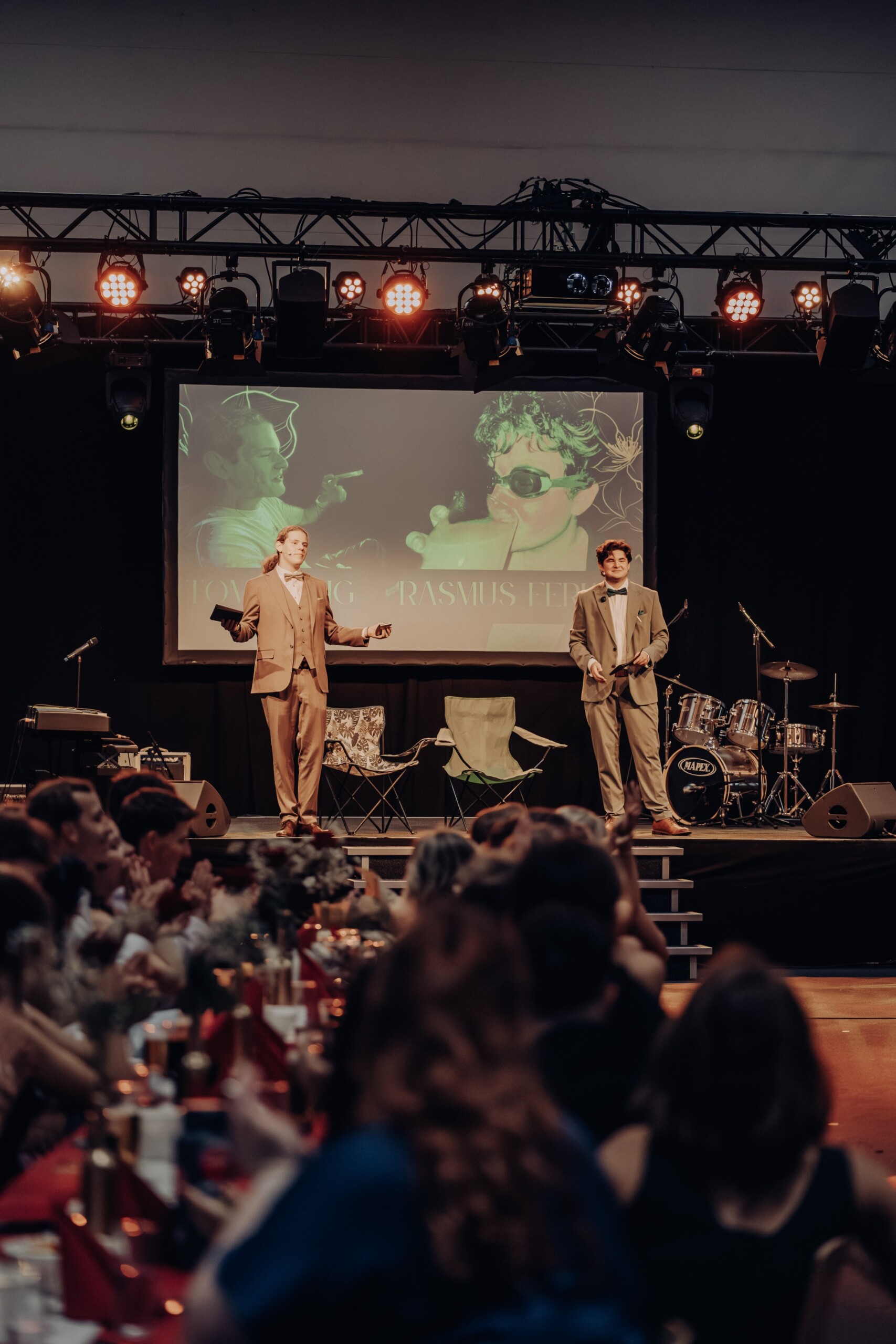 Tom Ohlig und Rasmus Ferko stehen als Moderatoren des Abiballs auf der Bühne – hinter ihnen eine projizierte Collage mit ihren Porträts, vor ihnen das applaudierende Publikum.