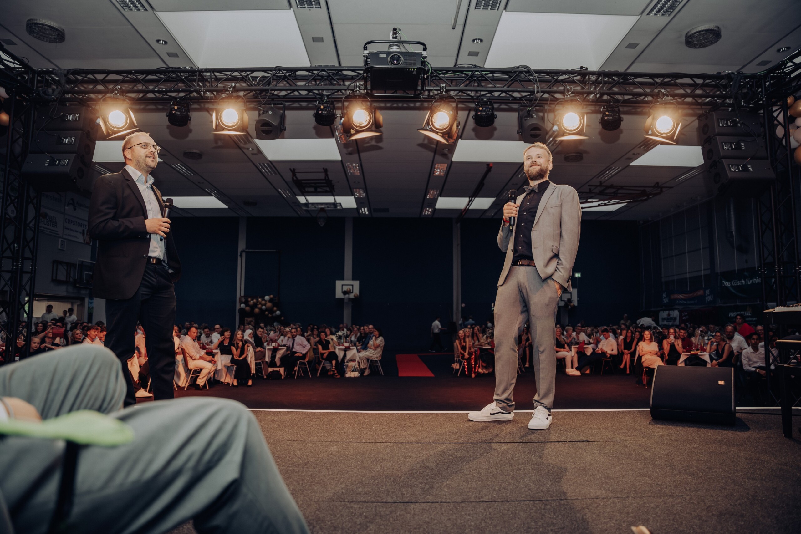 Sascha Stelzer und Florian Günther stehen mit Mikrofon auf der Bühne der Nutscheidhalle und gestalten humorvoll einen Programmpunkt – das Publikum sitzt an dekorierten Tischen.