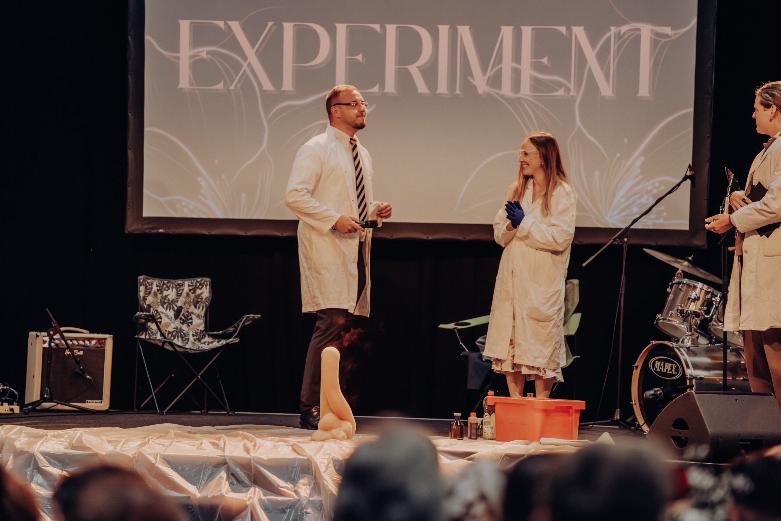 Lehrkräfte und Schüler:innen im Laborkittel präsentieren ein chemisches Bühnenerlebnis: Mitten im Experiment steigt ein Schaumberg empor – das Publikum ist gespannt.