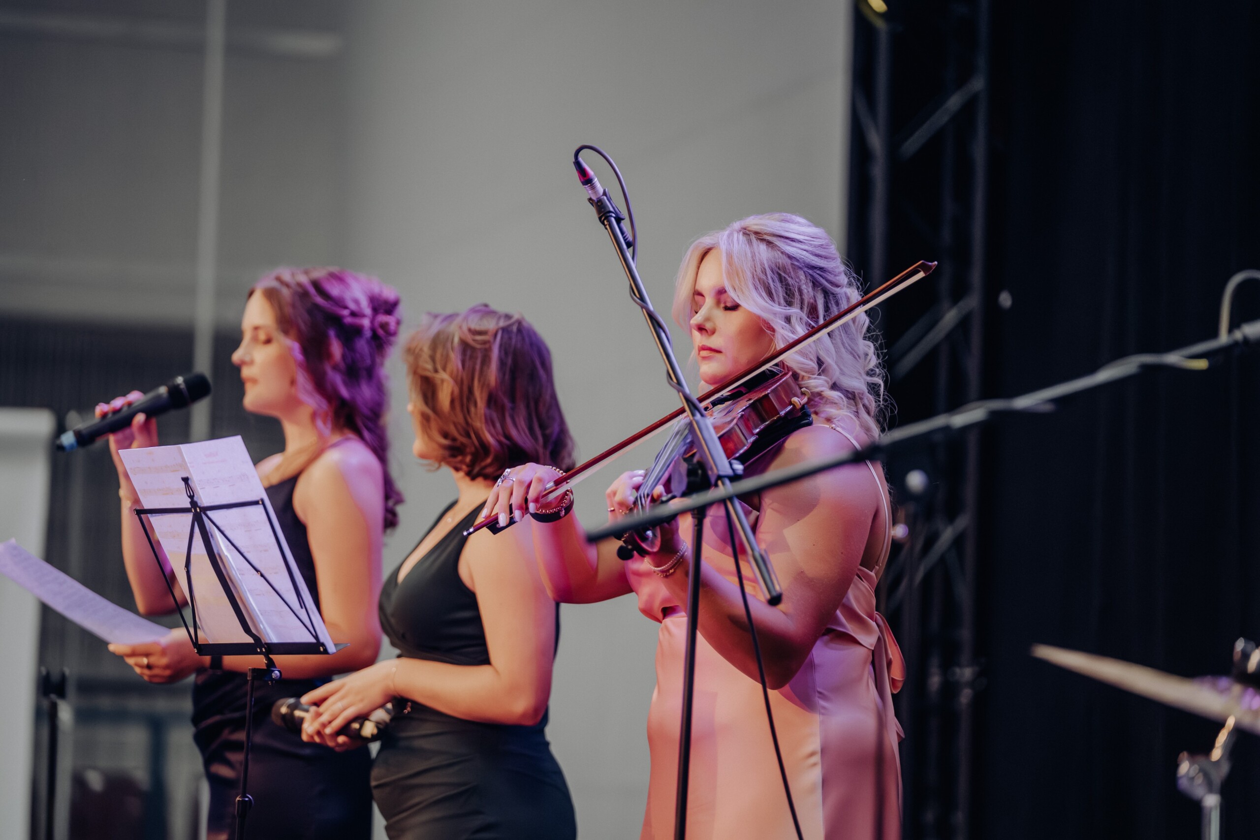 Zwei Abiturientinnen singen in Mikrofone, während eine dritte konzentriert Geige spielt. Alle drei stehen in festlicher Abendkleidung auf der Bühne, im Vordergrund ist ein Notenständer zu sehen..