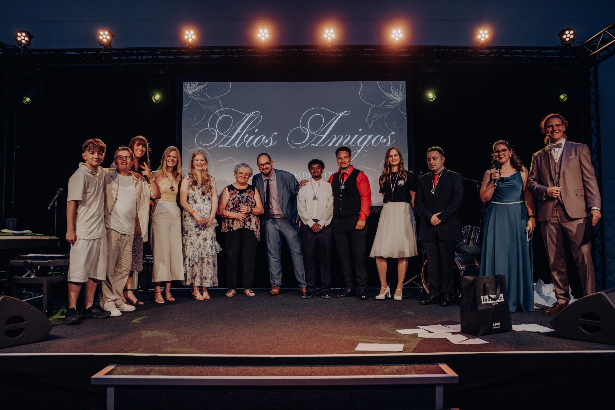 Eine Gruppe von Schüler:innen und Lehrkräften steht festlich gekleidet auf der Bühne, vor einem Bildschirm mit dem Schriftzug „Adios Amigos“. Sie posieren lächelnd für ein gemeinsames Erinnerungsfoto beim Abiball der Gesamtschule.