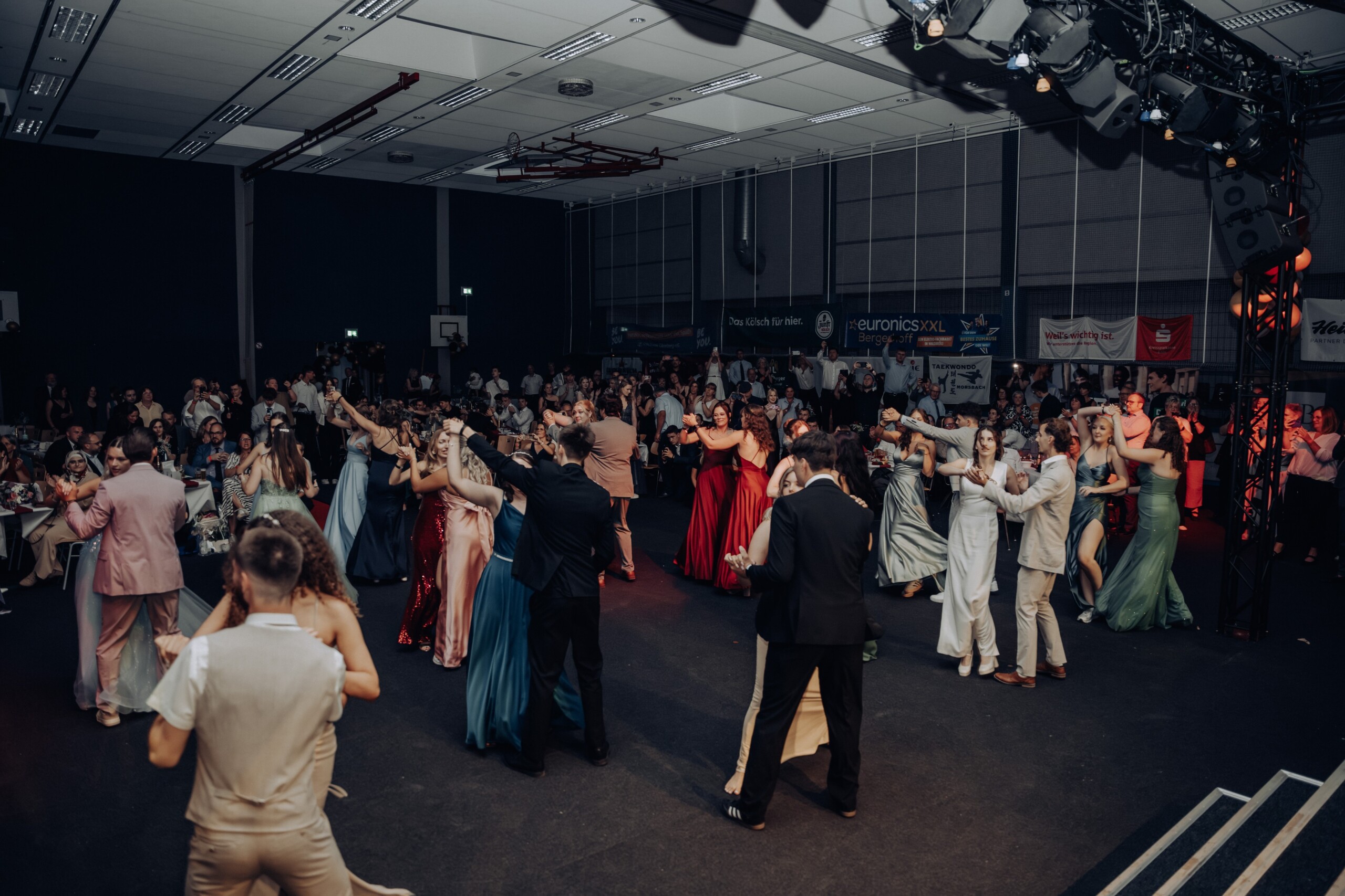 Zahlreiche Abiturient:innen tanzen in eleganter Abendgarderobe eng umschlungen auf der Tanzfläche. Im Hintergrund applaudieren und fotografieren Gäste, während eine feierliche Stimmung den Saal erfüllt.