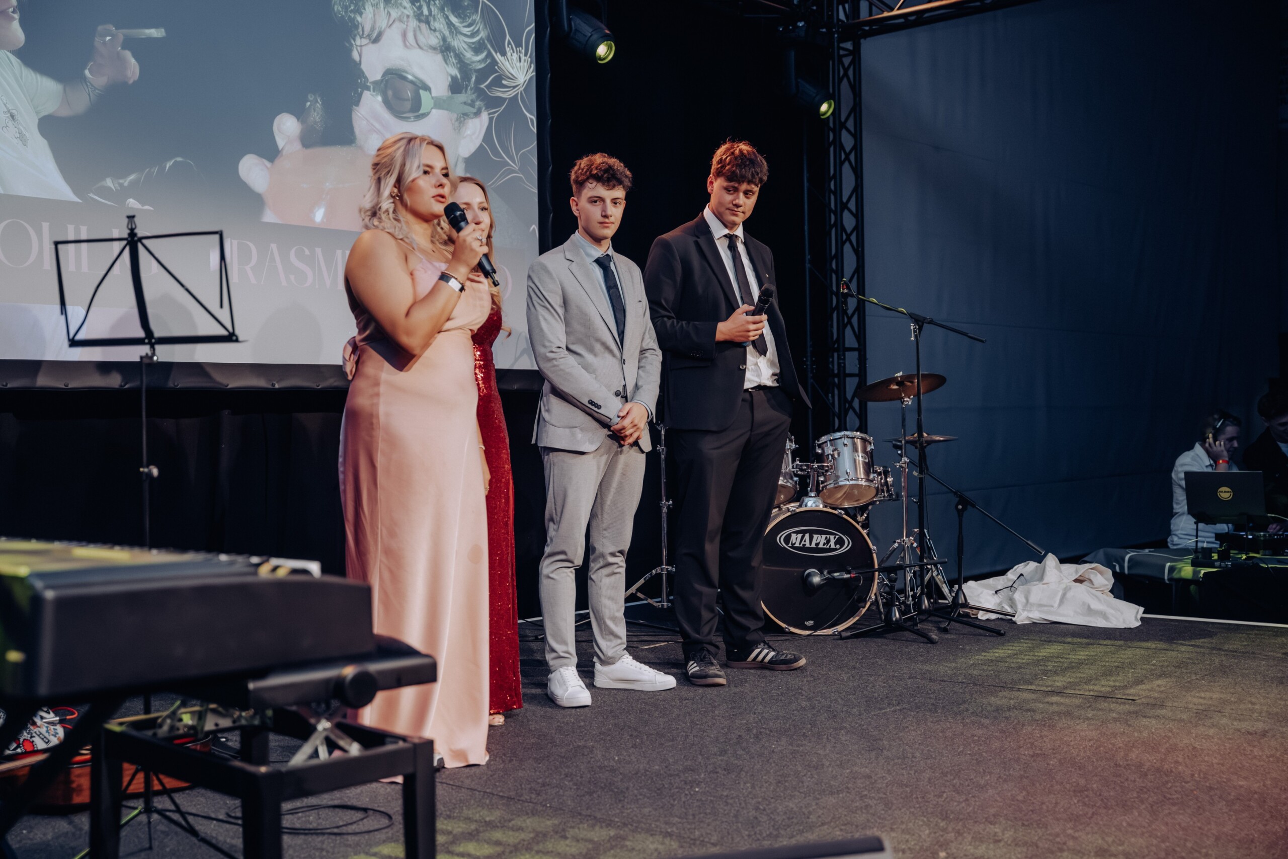 Vier Abiturient:innen stehen auf der Bühne, Mikrofone in der Hand. Sie blicken ernst und konzentriert ins Publikum, während hinter ihnen ein projiziertes Bild auf der Leinwand zu sehen ist.