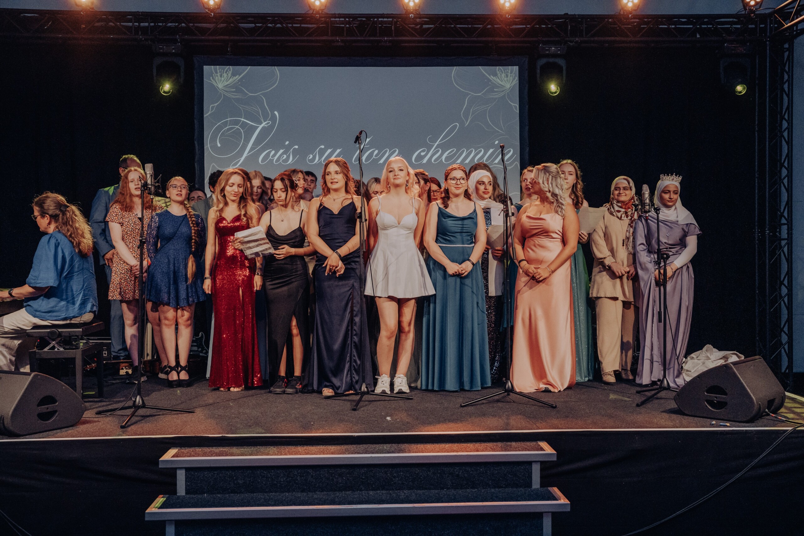 Ein großer Chor aus Abiturient:innen steht auf der Bühne und singt gemeinsam das Lied „Vois sur ton chemin“. Die Sänger:innen tragen festliche Abendkleidung, im Vordergrund ist eine Keyboardspielerin zu sehen.