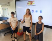 Drei strahlende Schüler:innen stehen vor einem digitalen Whiteboard mit dem Logo der Gesamtschule Waldbröl. In der Mitte hält eine Schülerin den Eisgutschein in der Hand, überreicht von den beiden SV-Initiatorinnen des Fahrradwettbewerbs.
