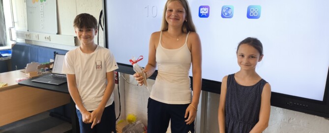 Drei strahlende Schüler:innen stehen vor einem digitalen Whiteboard mit dem Logo der Gesamtschule Waldbröl. In der Mitte hält eine Schülerin den Eisgutschein in der Hand, überreicht von den beiden SV-Initiatorinnen des Fahrradwettbewerbs.