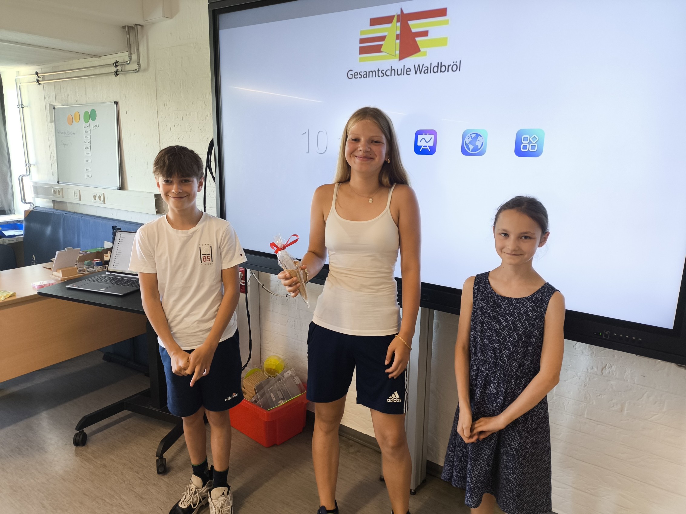Drei strahlende Schüler:innen stehen vor einem digitalen Whiteboard mit dem Logo der Gesamtschule Waldbröl. In der Mitte hält eine Schülerin den Eisgutschein in der Hand, überreicht von den beiden SV-Initiatorinnen des Fahrradwettbewerbs.