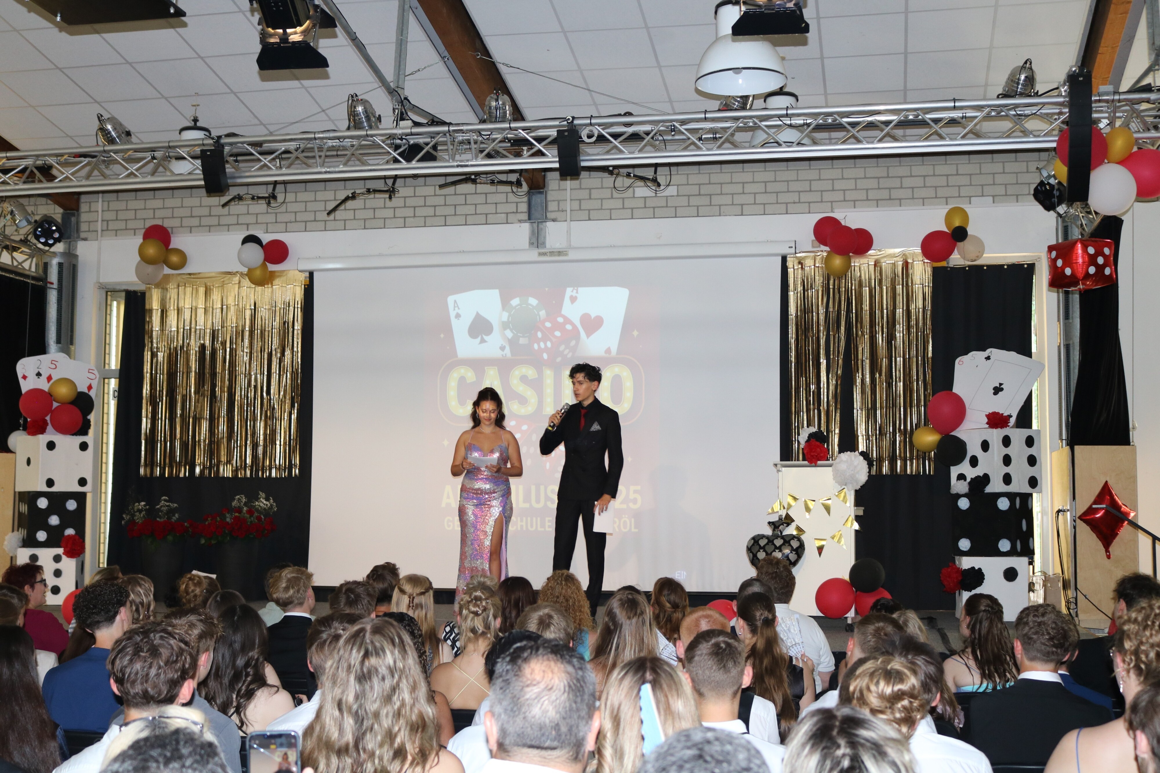 Zwei weitere Moderator:innen präsentieren auf der Bühne vor voll besetztem Saal, flankiert von selbst gebastelter Casino-Deko mit Würfeln, Karten und Ballons.