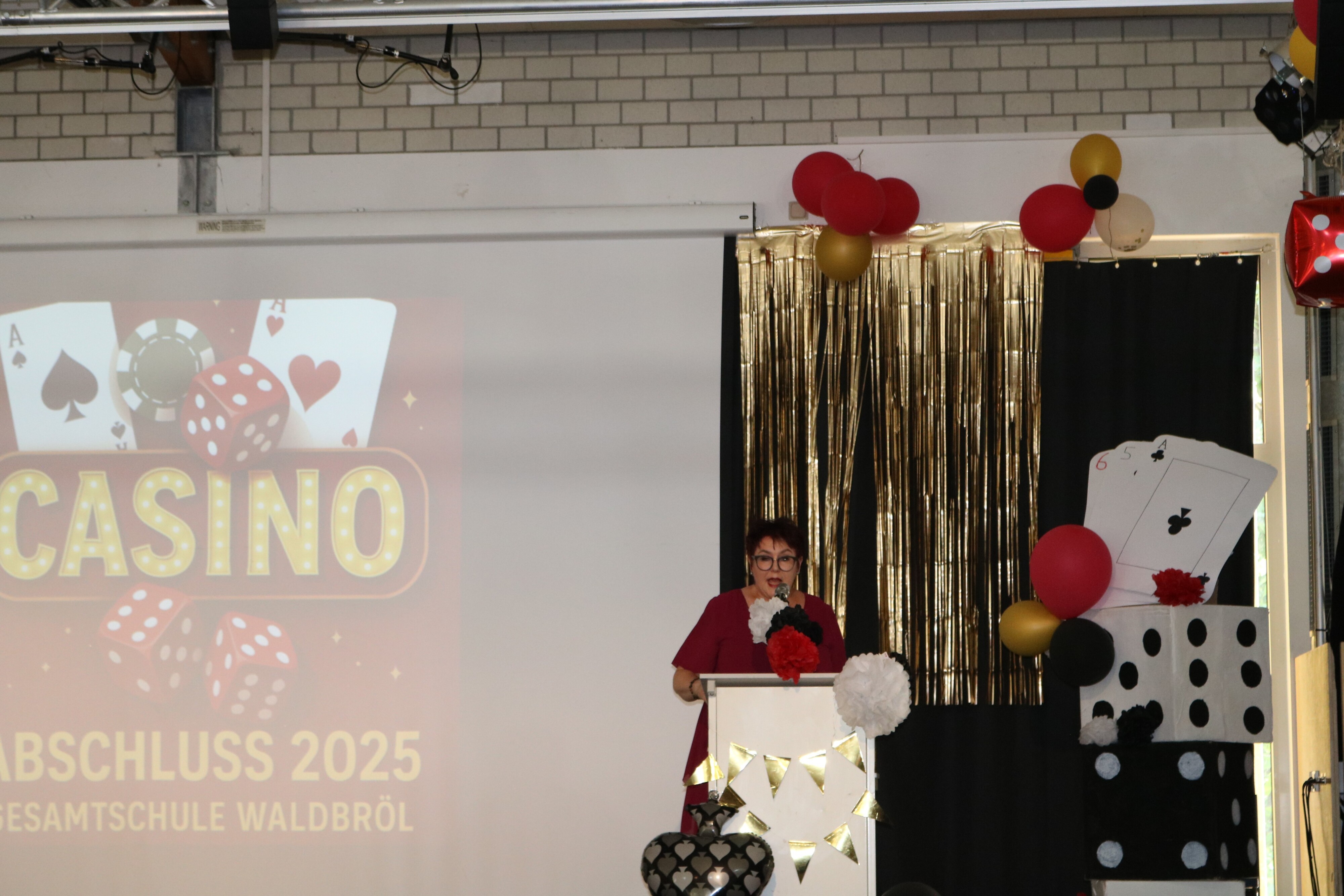 Schulleiterin Kirsten Wallbaum hält ihre Rede am Rednerpult, umgeben von aufwändiger Casino-Dekoration mit Spielkarten und Girlanden.