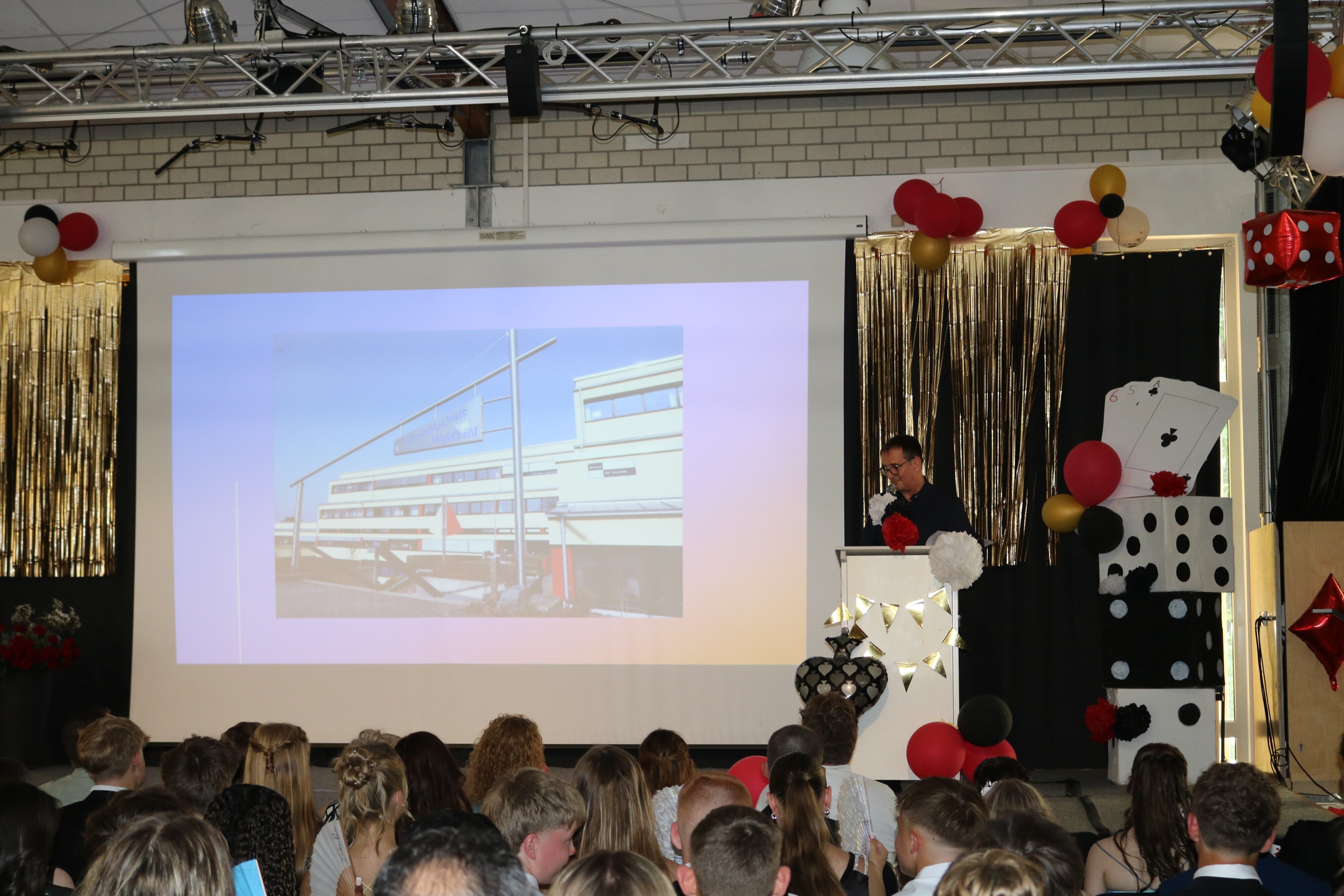Paul Giebeler bei seinem spontanen Grußwort am Pult, im Hintergrund ein Foto der Schule auf der Leinwand, davor das gespannte Publikum.