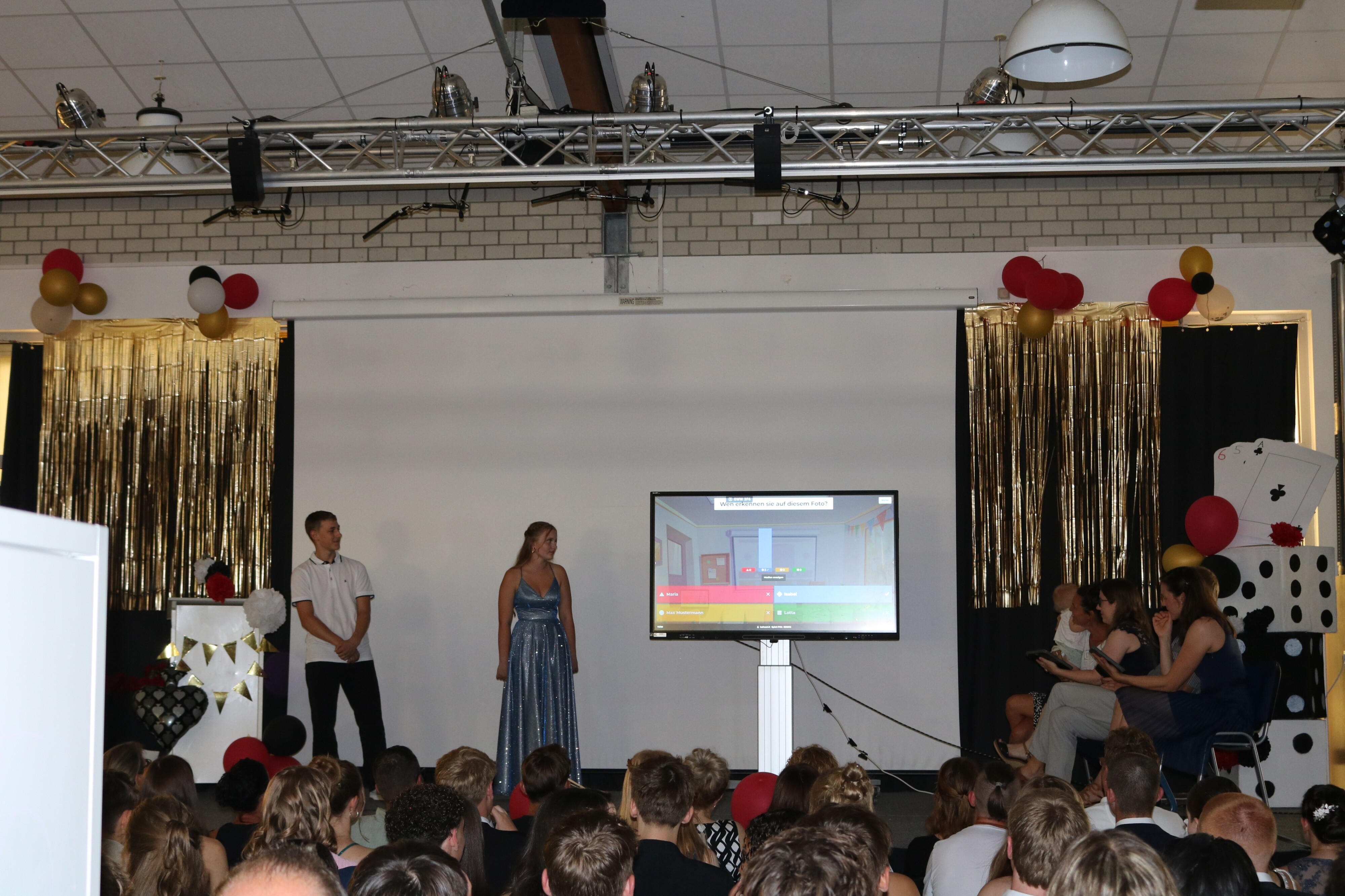 Zwei Schüler:innen stehen auf der Bühne, daneben ein großer Monitor mit einer Kahoot-Fragerunde. Das Publikum folgt gespannt dem Quiz.