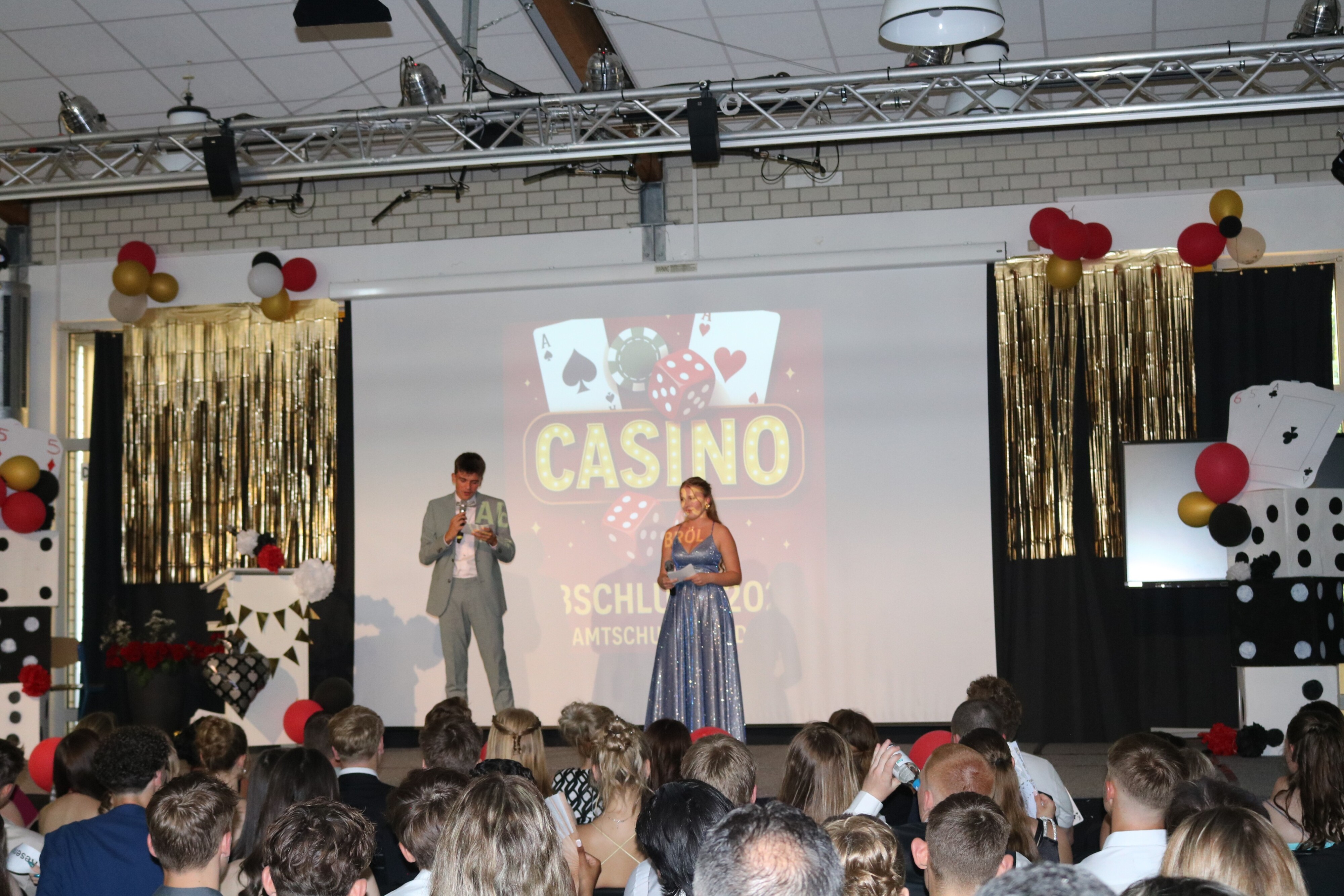 Das Moderationsteam eröffnet die Abschlussfeier, im Vordergrund das Publikum, im Hintergrund die große Motto-Projektion „Casino – Abschluss 2025“.