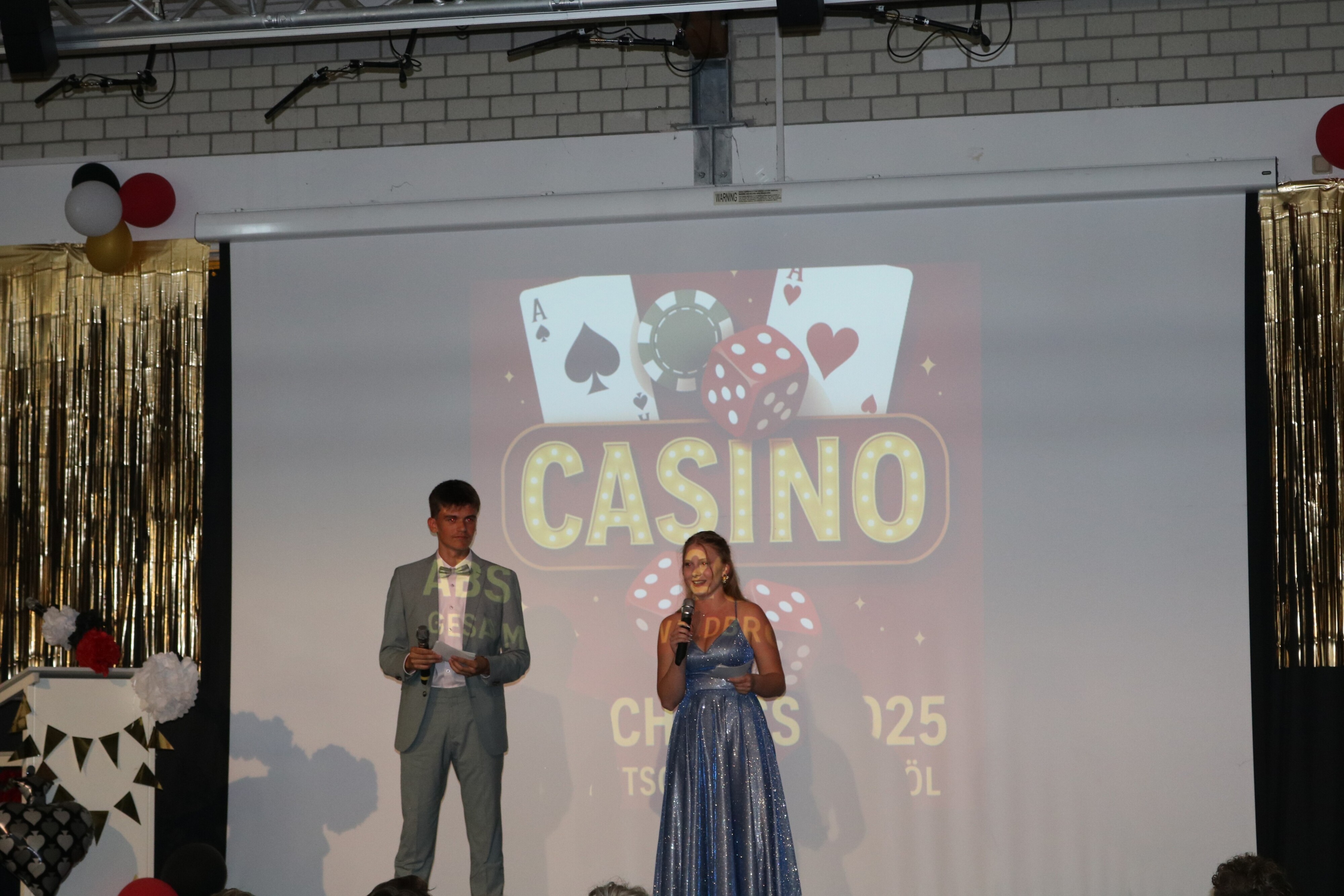 Das Moderationsteam eröffnet die Abschlussfeier, im Vordergrund das Publikum, im Hintergrund die große Motto-Projektion „Casino – Abschluss 2025“.
