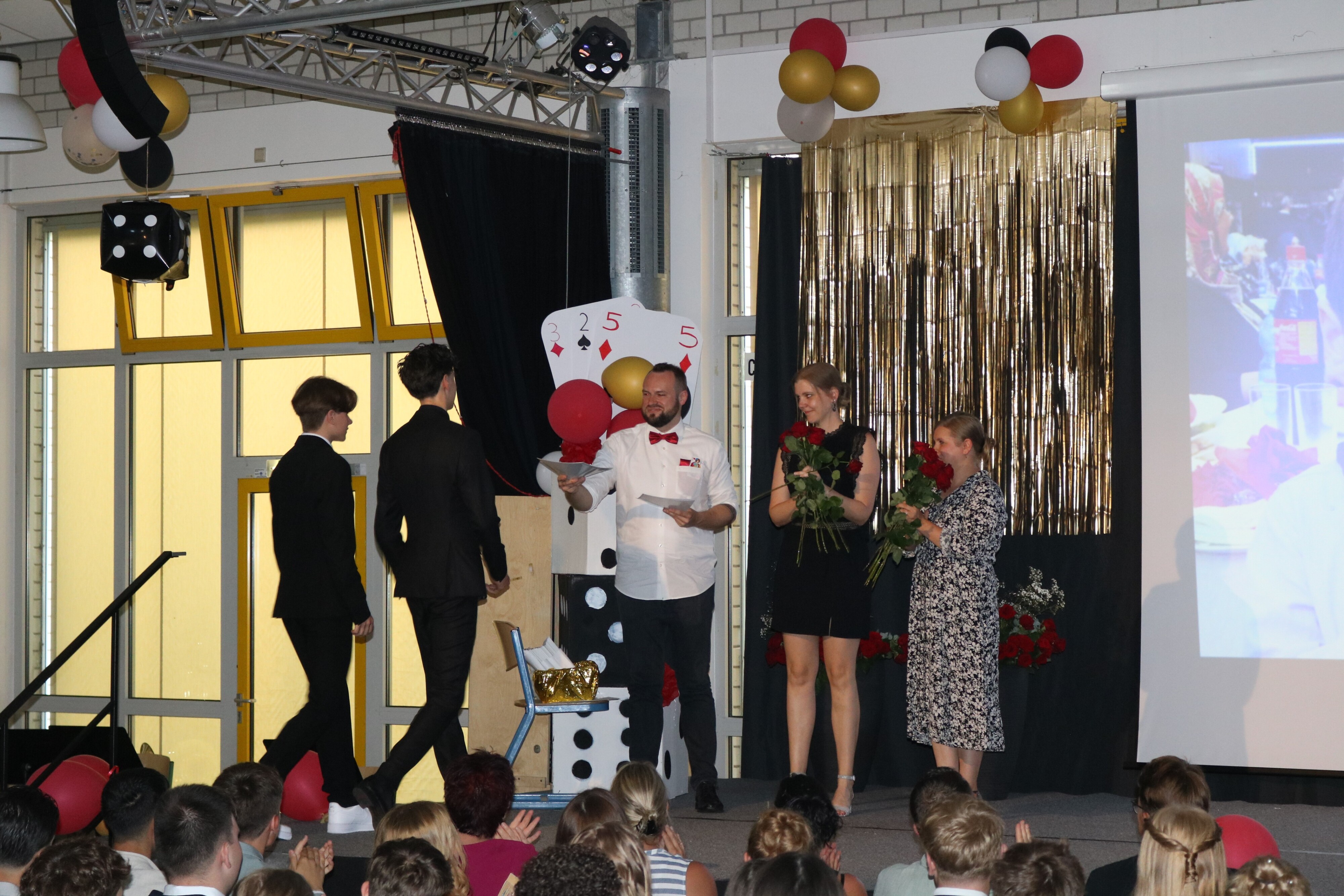 Zwei Schüler betreten die Bühne zur Zeugnisvergabe, wo sie von drei Lehrkräften mit Urkunden und Rosen empfangen werden.
