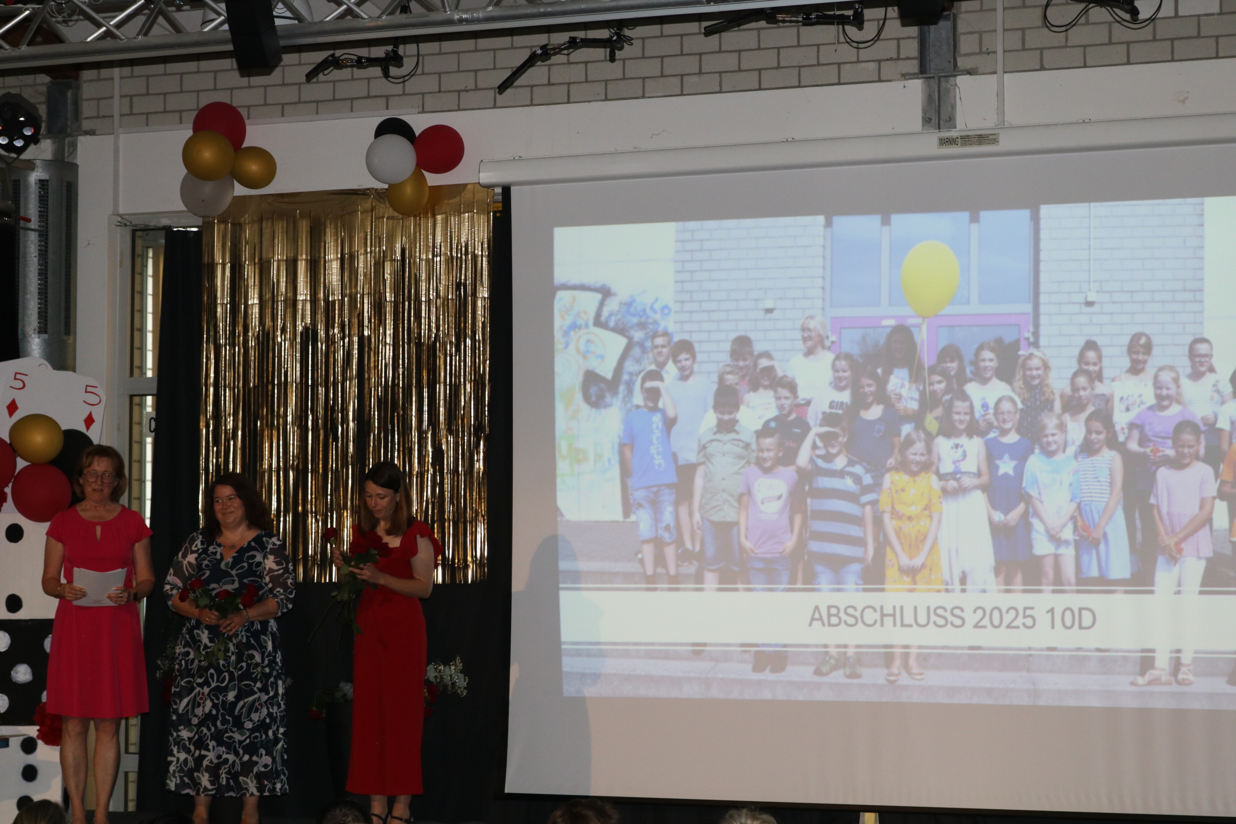 Drei Lehrerinnen präsentieren die Abschlussklasse 10d, während im Hintergrund ein altes Klassenfoto auf der Leinwand erscheint.