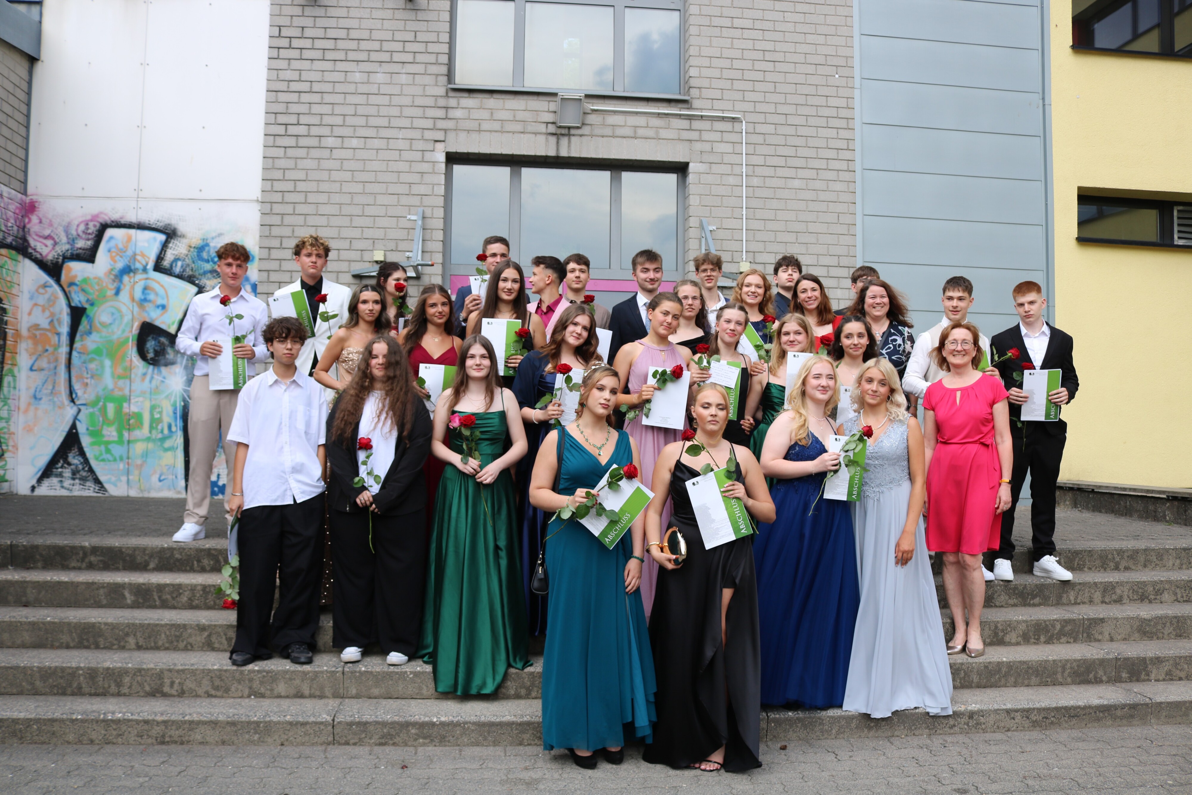 Die Klasse 10d posiert mit Urkunden und Rosen für ein Gruppenfoto auf der Freitreppe vor dem Schulgebäude – stolze Gesichter und festliche Kleidung prägen das Bild.