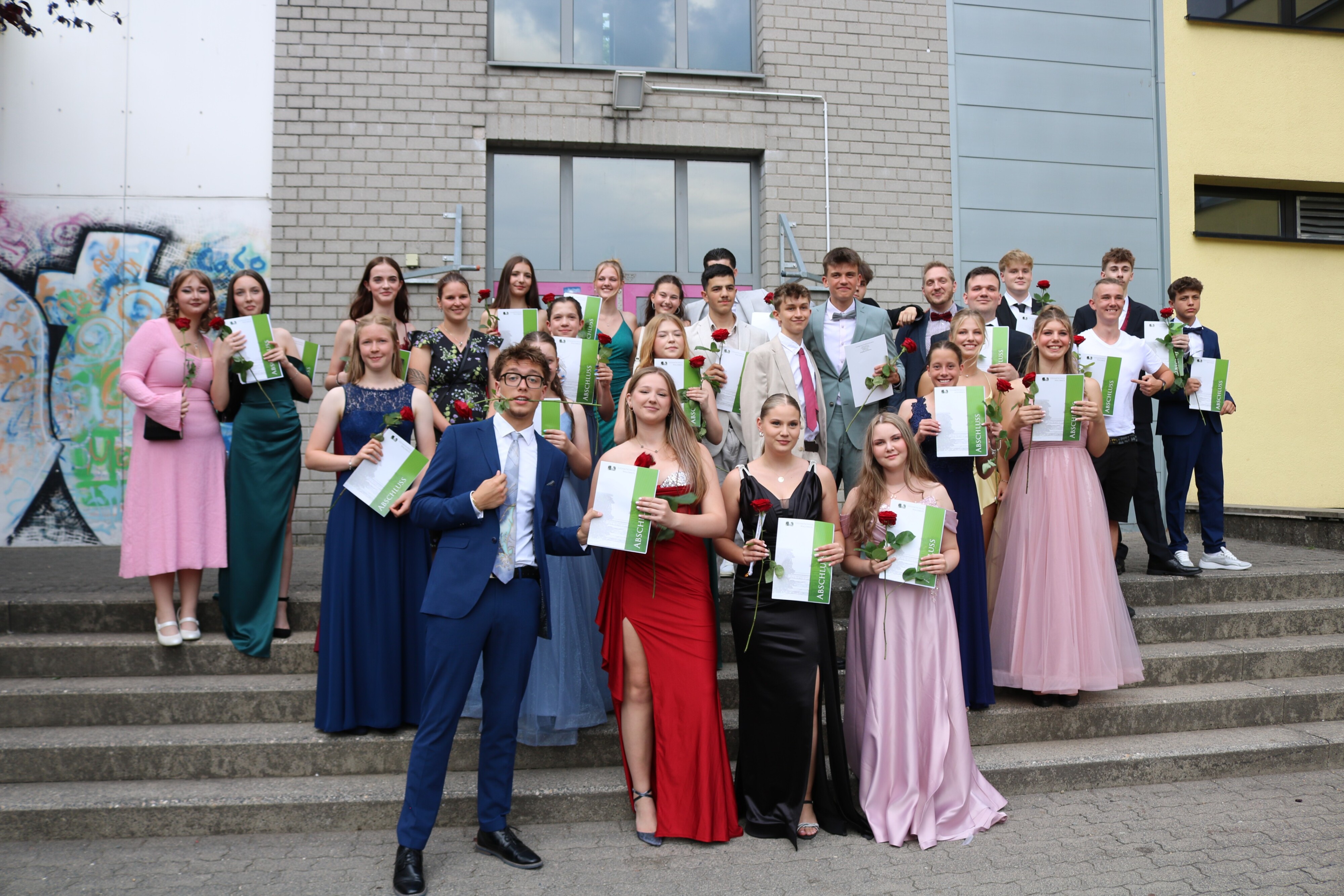Gruppenfoto einer weiteren Abschlussklasse auf der Freitreppe vor der Schule – Urkunden und Rosen in den Händen, festliche Stimmung im Bild eingefangen.
