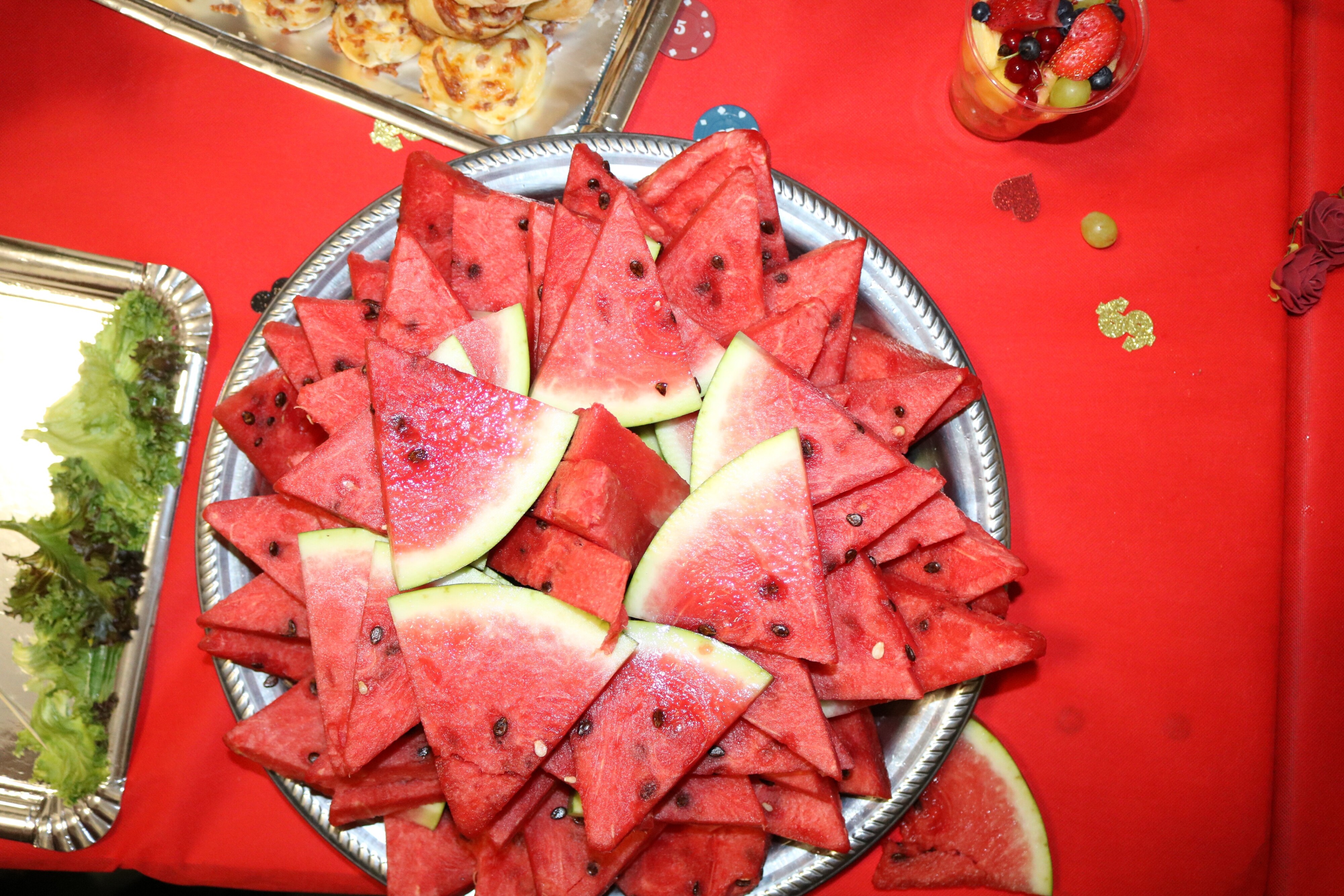 Frische Wassermelone in Dreiecks-Stücken auf einem silbernen Tablett – sommerlich angerichtet, umgeben von rotem Tischtuch und Dekokonfetti in Pokerchips-Optik.
