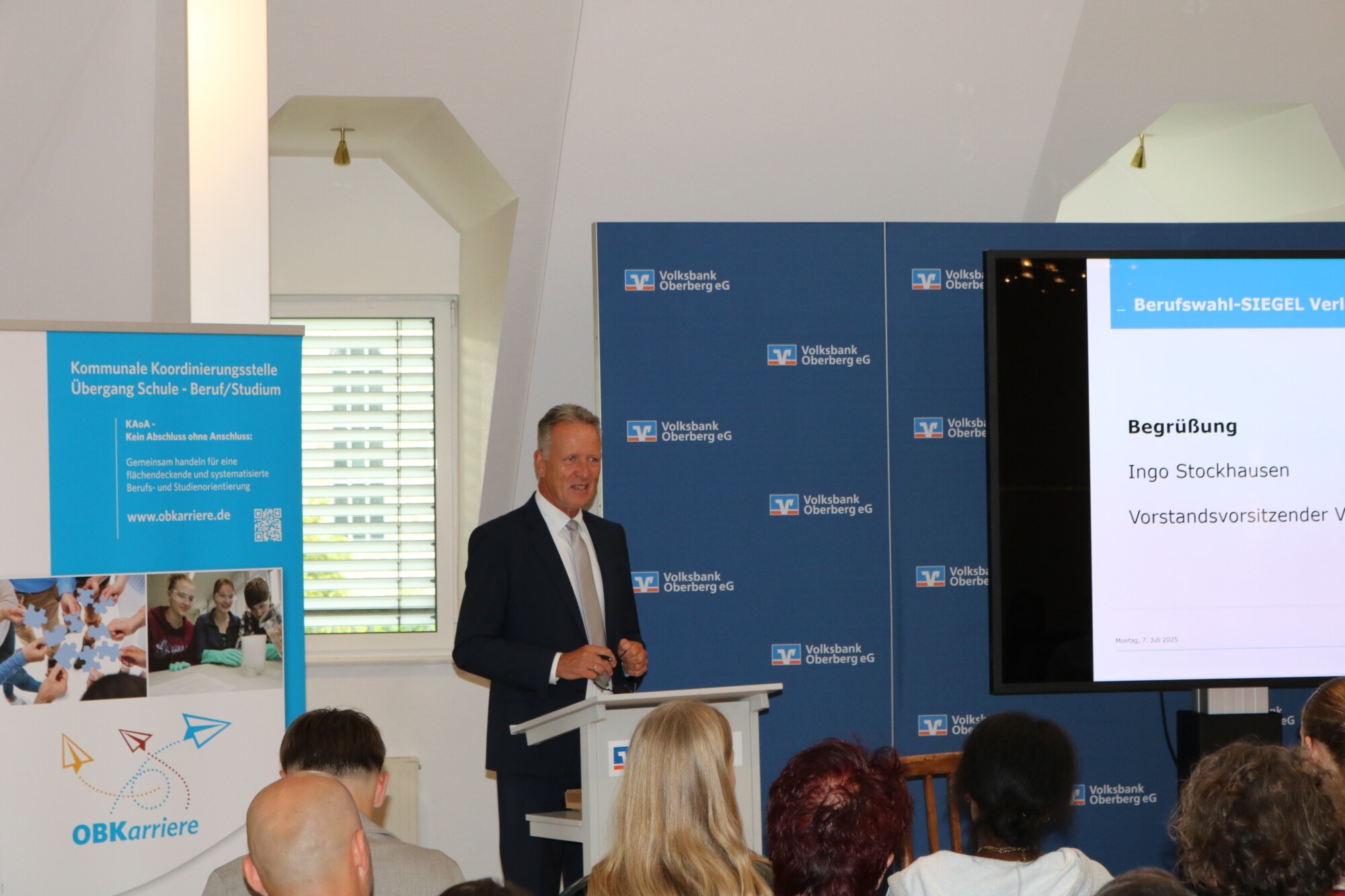 Begrüßung durch Ingo Stockhausen, Vorstandsvorsitzender der Volksbank Oberberg, bei der Verleihung des Berufswahl-Siegels 2025. Im Hintergrund Banner der kommunalen Koordinierungsstelle und Logos der Volksbank.
