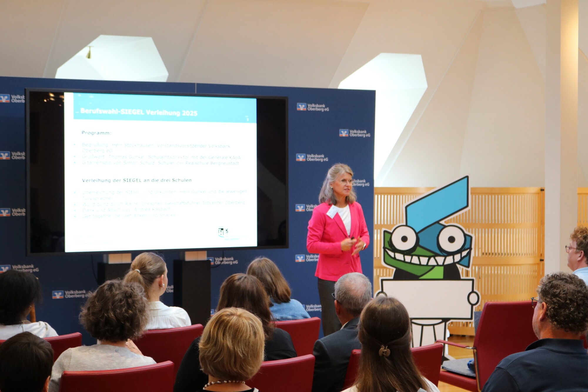 Moderatorin der Verleihungsveranstaltung des Berufswahl-Siegels 2025 spricht vor Publikum. Auf der Präsentationsfolie sind Programmpunkte und der stilisierte Siegel-Roboter zu sehen.