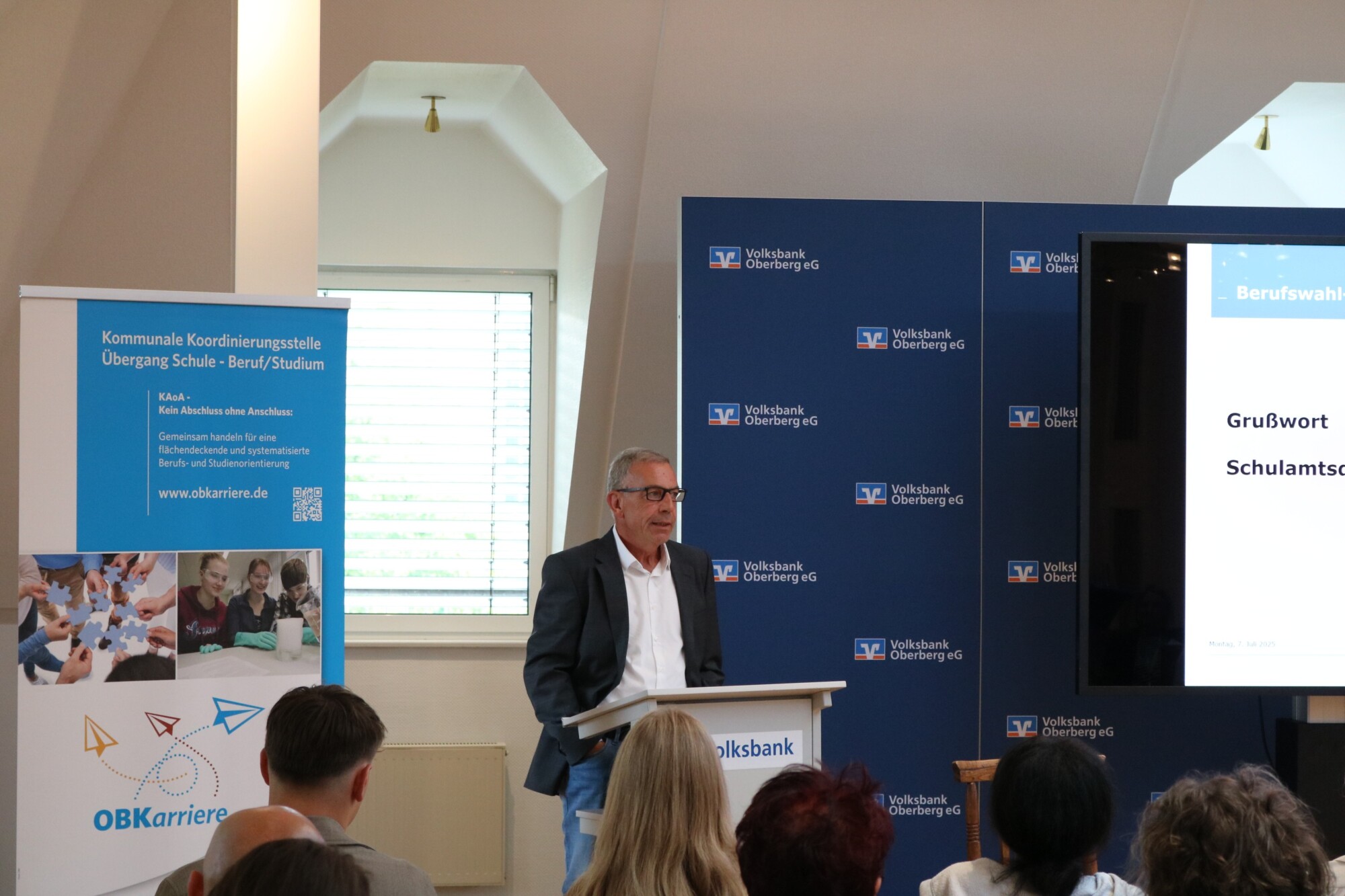 Grußwort durch einen Vertreter des Schulamts auf der Bühne der Volksbank Oberberg. Im Hintergrund ein Roll-Up der kommunalen Koordinierungsstelle.