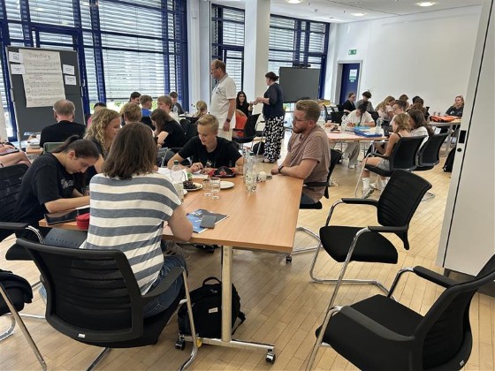 Blick in den gut besuchten Veranstaltungsraum: An mehreren Tischen sitzen Schülerinnen und Schüler in Gruppen und arbeiten gemeinsam an ihren Projekten.