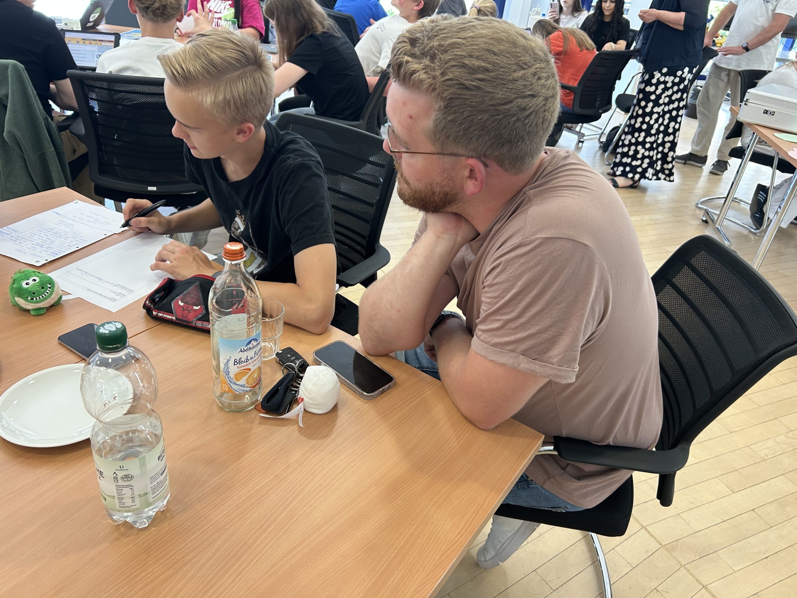 Ein Jugendlicher schreibt Notizen auf ein Arbeitsblatt, neben ihm sitzt ein Betreuer; auf dem Tisch stehen Getränke, Smartphones und kleine Maskottchen.