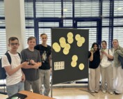 Die Gruppe der Social Media Scouts steht vor einer Präsentationstafel mit Stichworten zum Thema Medienkompetenz; sie halten kleine Maskottchen in den Händen.