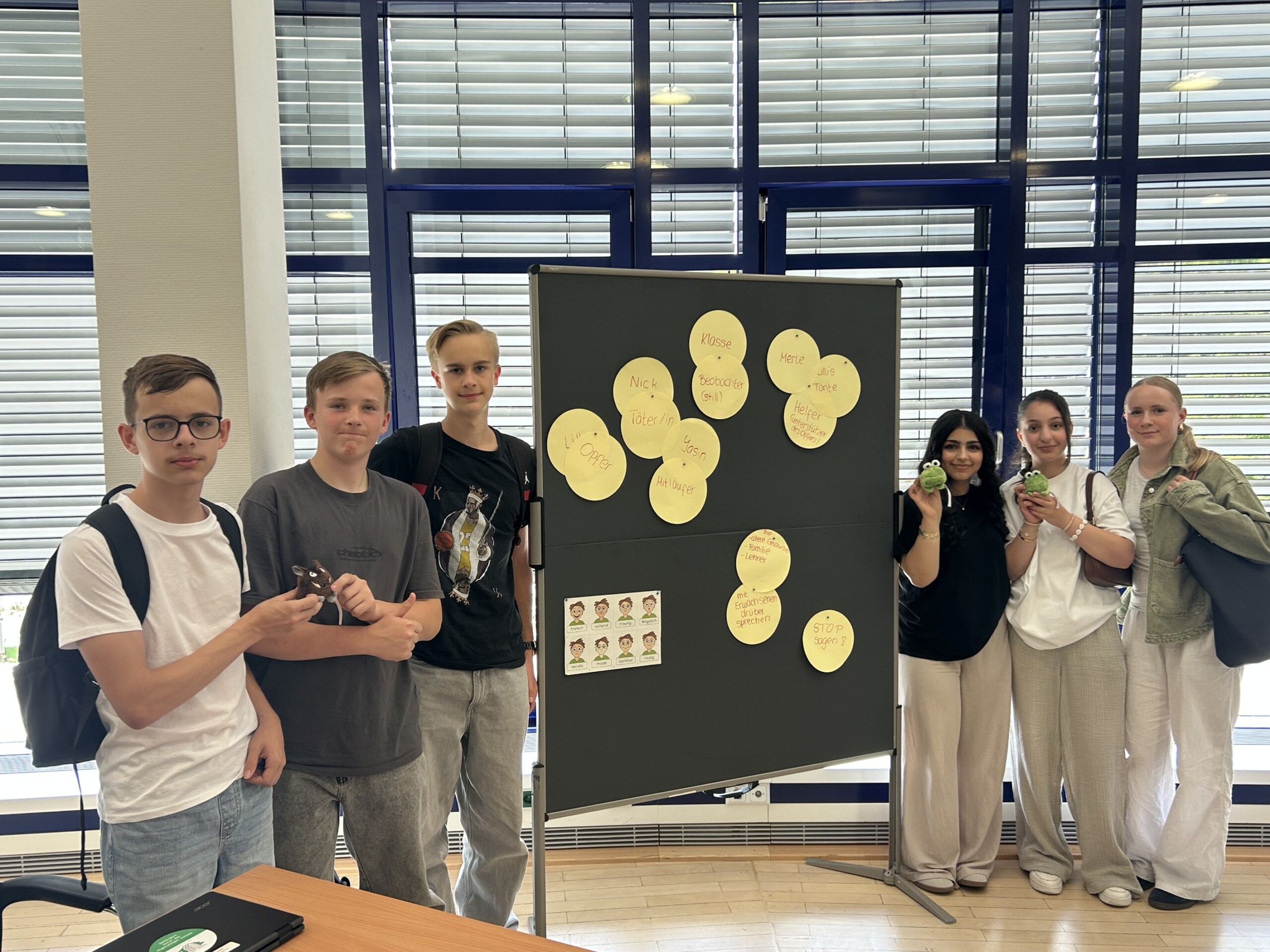 Die Gruppe der Social Media Scouts steht vor einer Präsentationstafel mit Stichworten zum Thema Medienkompetenz; sie halten kleine Maskottchen in den Händen.