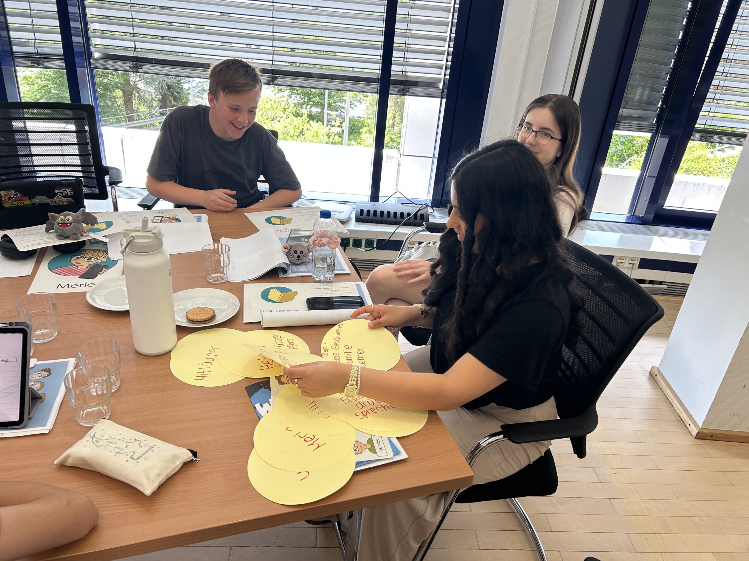 Drei Jugendliche arbeiten konzentriert an einem Tisch voller Materialien und Snacks; sie gestalten farbige Kreise mit Begriffen zum Thema Social Media.