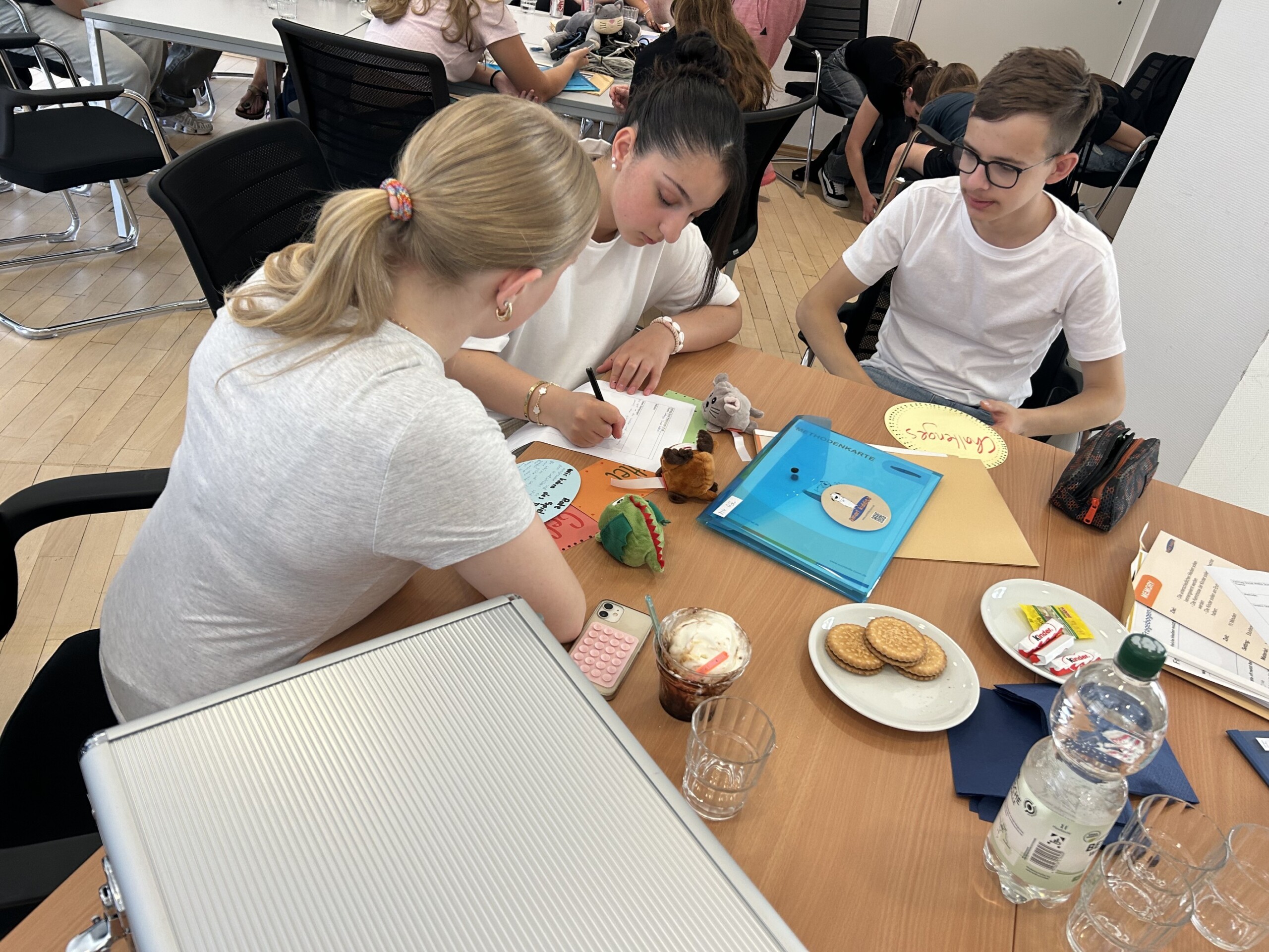 Zwei Mädchen und ein Junge sitzen an einem Tisch und arbeiten mit großen, gelben Moderationskarten; auf dem Tisch liegen Unterlagen, Getränke und Plüschfiguren.