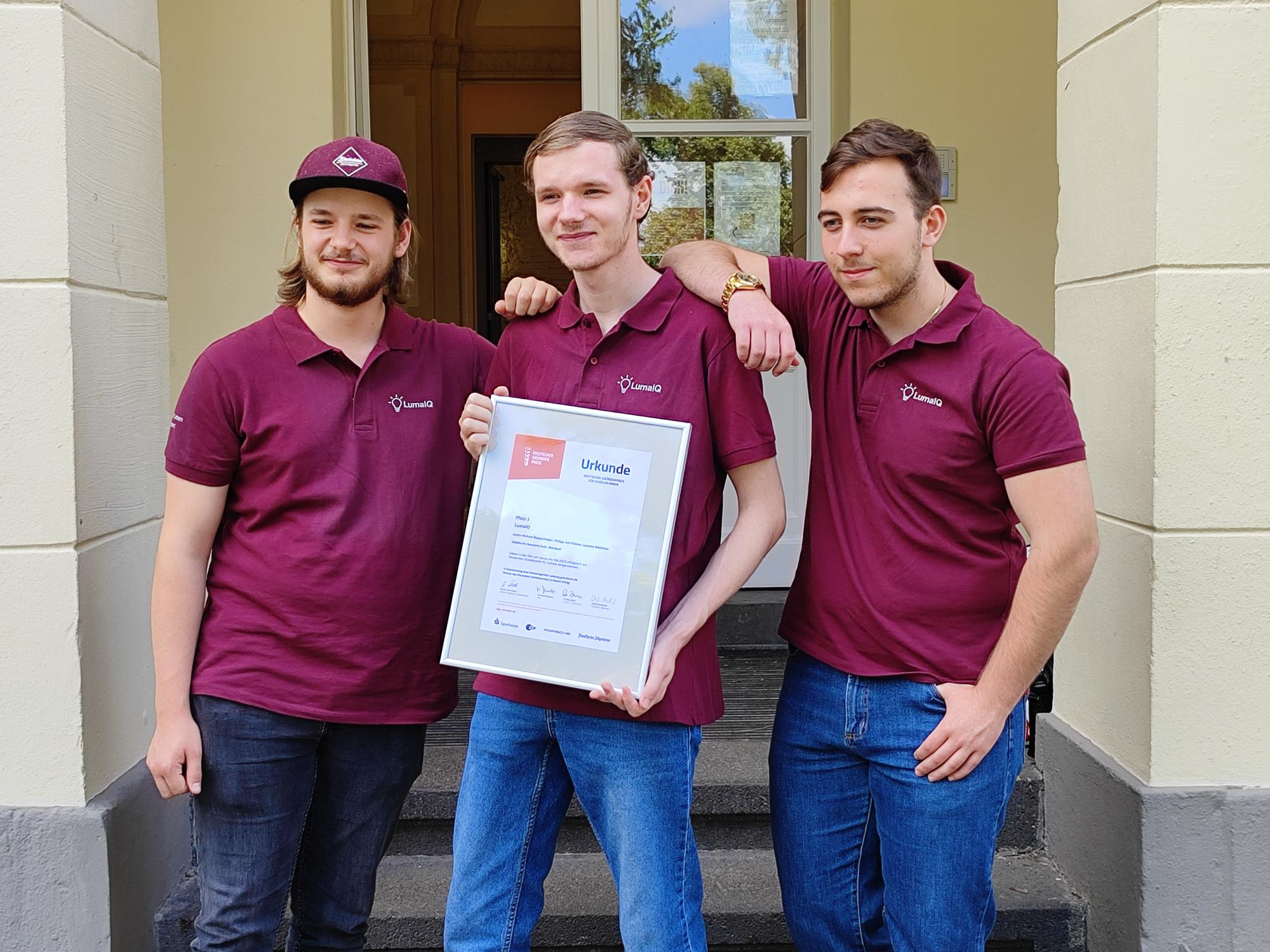 Drei Mitglieder des Teams LumaIQ auf der Treppe des Schlosses: In einheitlichen Polo-Shirts präsentieren sie die Urkunde für den 5. Platz, alle drei lächeln selbstbewusst in die Kamera.
