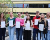 Acht Schüler:innen des Cambridge-Projektkurses der Gesamtschule Waldbröl halten stolz ihre Cambridge-Zertifikate in den Händen, gemeinsam mit Kursleiterin Lisa Dornseifer.