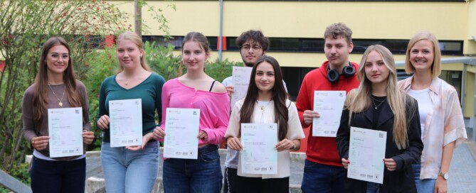 Acht Schüler:innen des Cambridge-Projektkurses der Gesamtschule Waldbröl halten stolz ihre Cambridge-Zertifikate in den Händen, gemeinsam mit Kursleiterin Lisa Dornseifer.