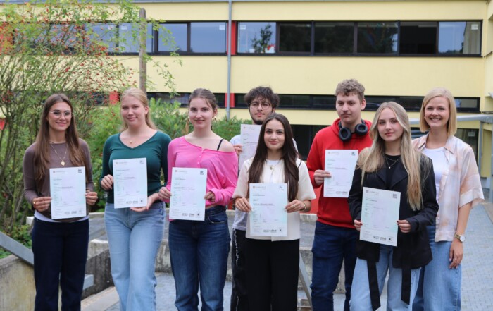 Acht Schüler:innen des Cambridge-Projektkurses der Gesamtschule Waldbröl halten stolz ihre Cambridge-Zertifikate in den Händen, gemeinsam mit Kursleiterin Lisa Dornseifer.