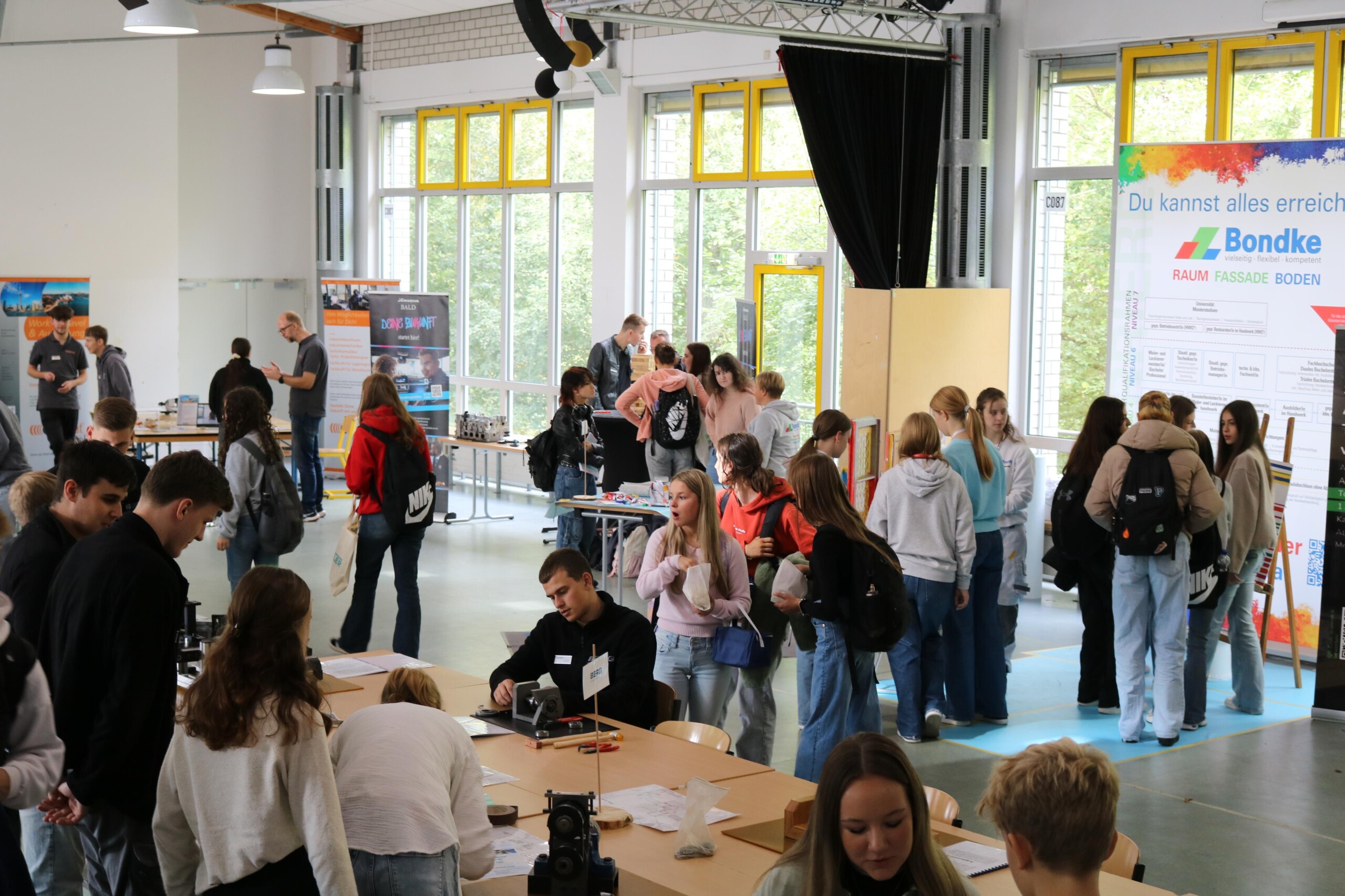 Schülerinnen und Schüler stehen in einer Schulmensa an verschiedenen Ständen, informieren sich und führen Gespräche.