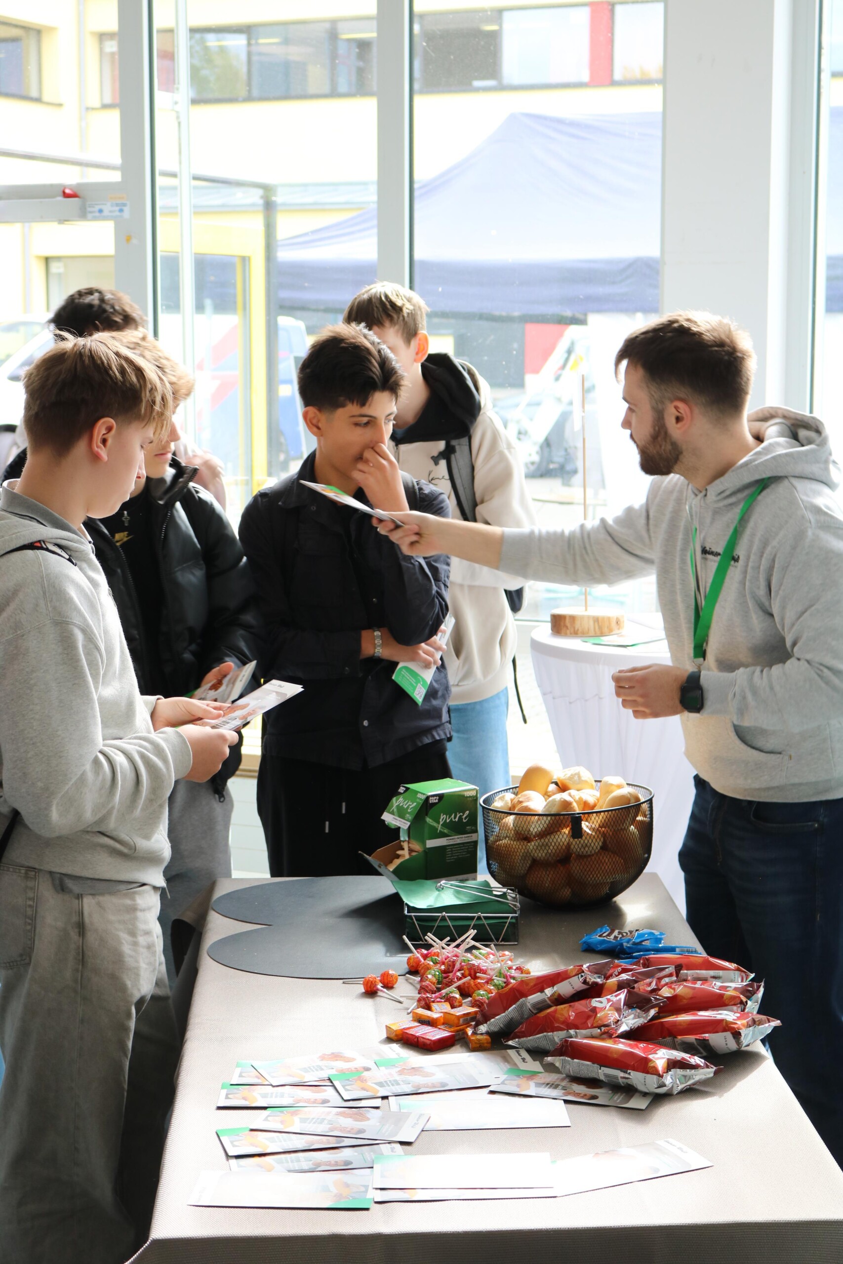 Eine Gruppe von Jungen steht an einem Stand, auf dessen Tisch Lebensmittelproben und Informationsmaterialien ausliegen. Ein Mann erklärt etwas und reicht Broschüren.