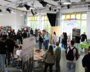 Viele Schülerinnen und Schüler bewegen sich durch einen großen Raum mit zahlreichen Infoständen. Gespräche und Begegnungen prägen die Atmosphäre.
