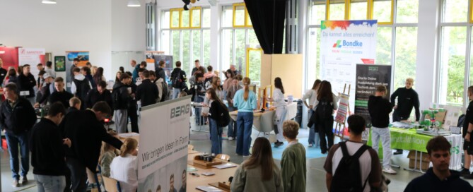 Viele Schülerinnen und Schüler bewegen sich durch einen großen Raum mit zahlreichen Infoständen. Gespräche und Begegnungen prägen die Atmosphäre.