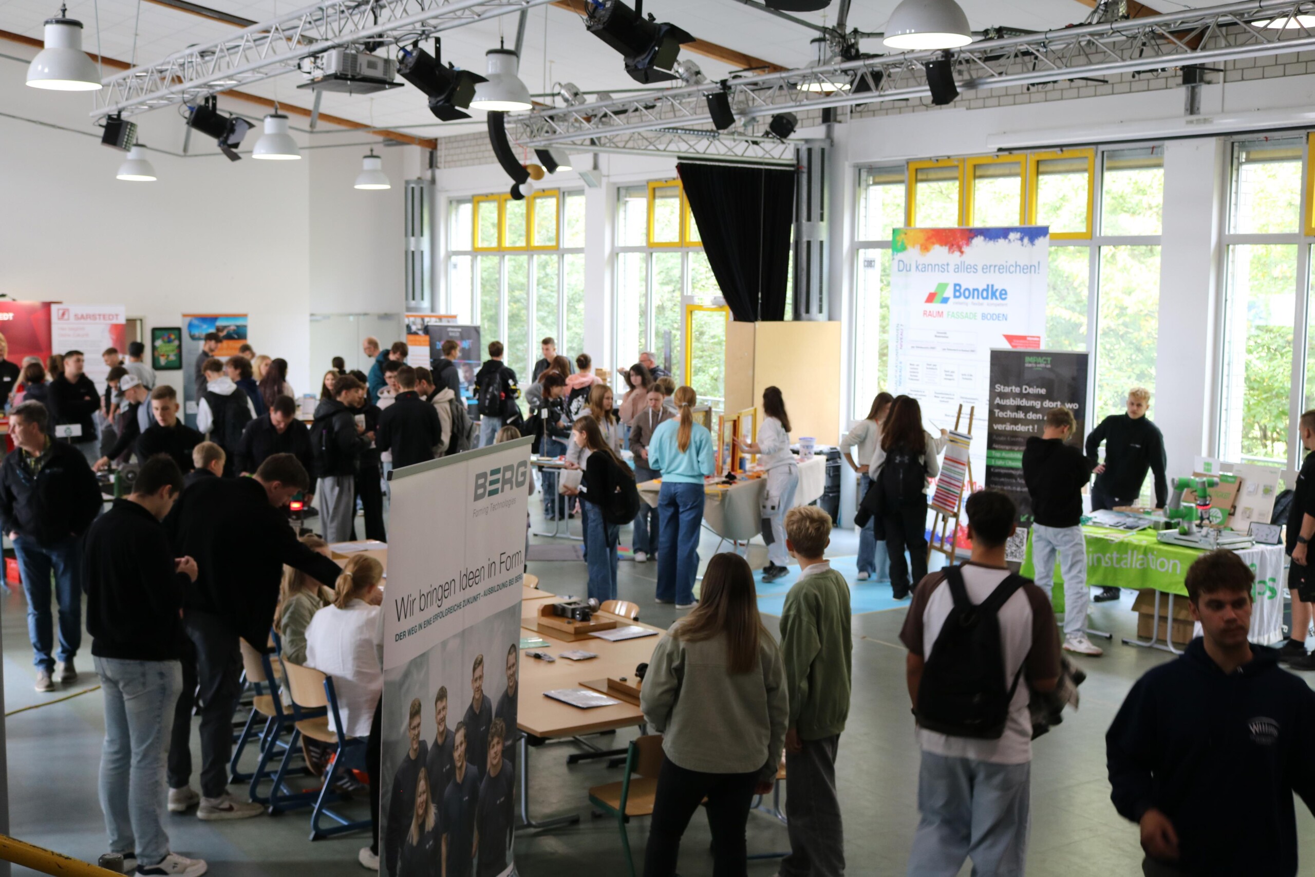 Viele Schülerinnen und Schüler bewegen sich durch einen großen Raum mit zahlreichen Infoständen. Gespräche und Begegnungen prägen die Atmosphäre.